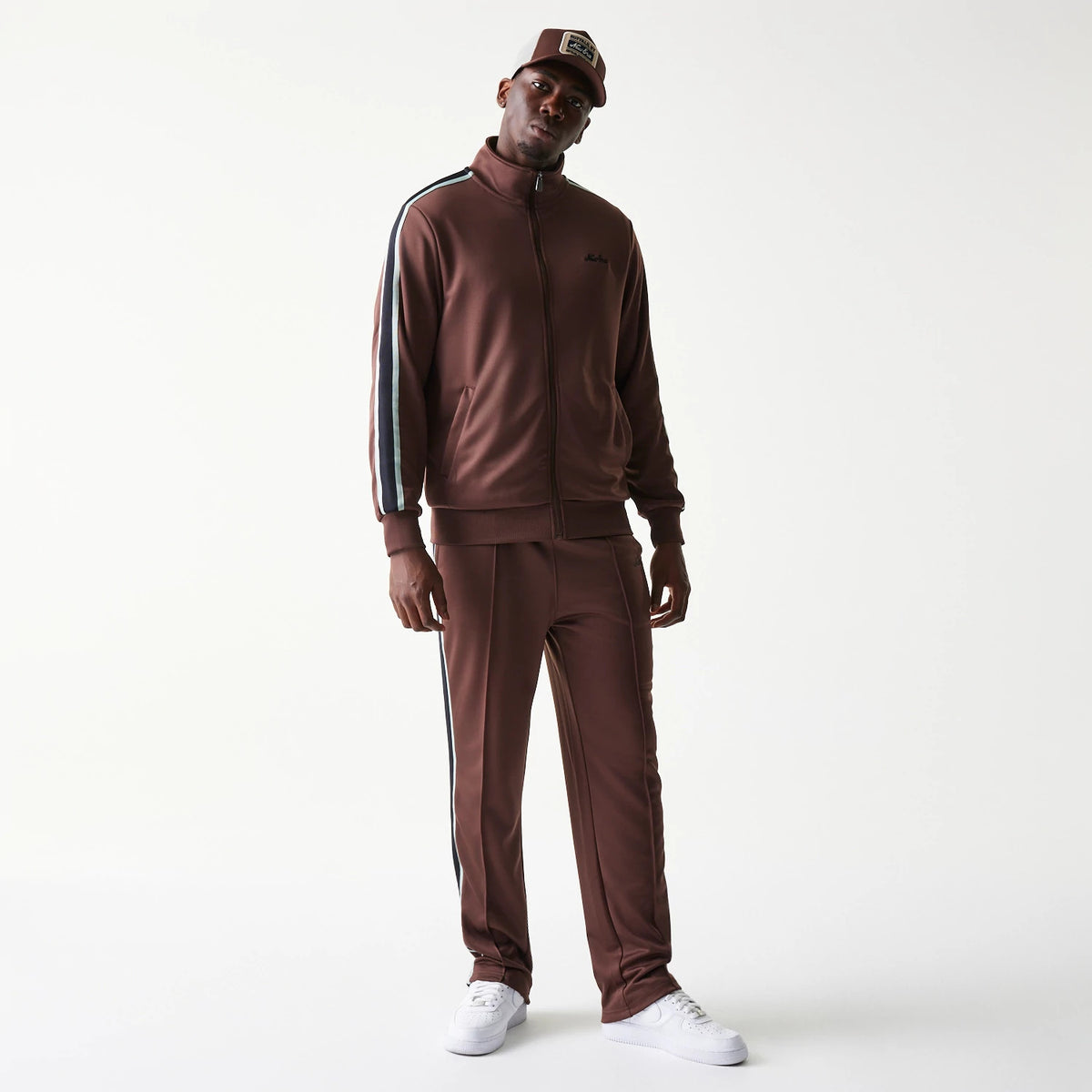 New Era Script Brown Straight Leg Joggers, on-model front view showing the relaxed fit with white and black stripe along the sides, paired with white trainers and a matching brown zip-up jacket.