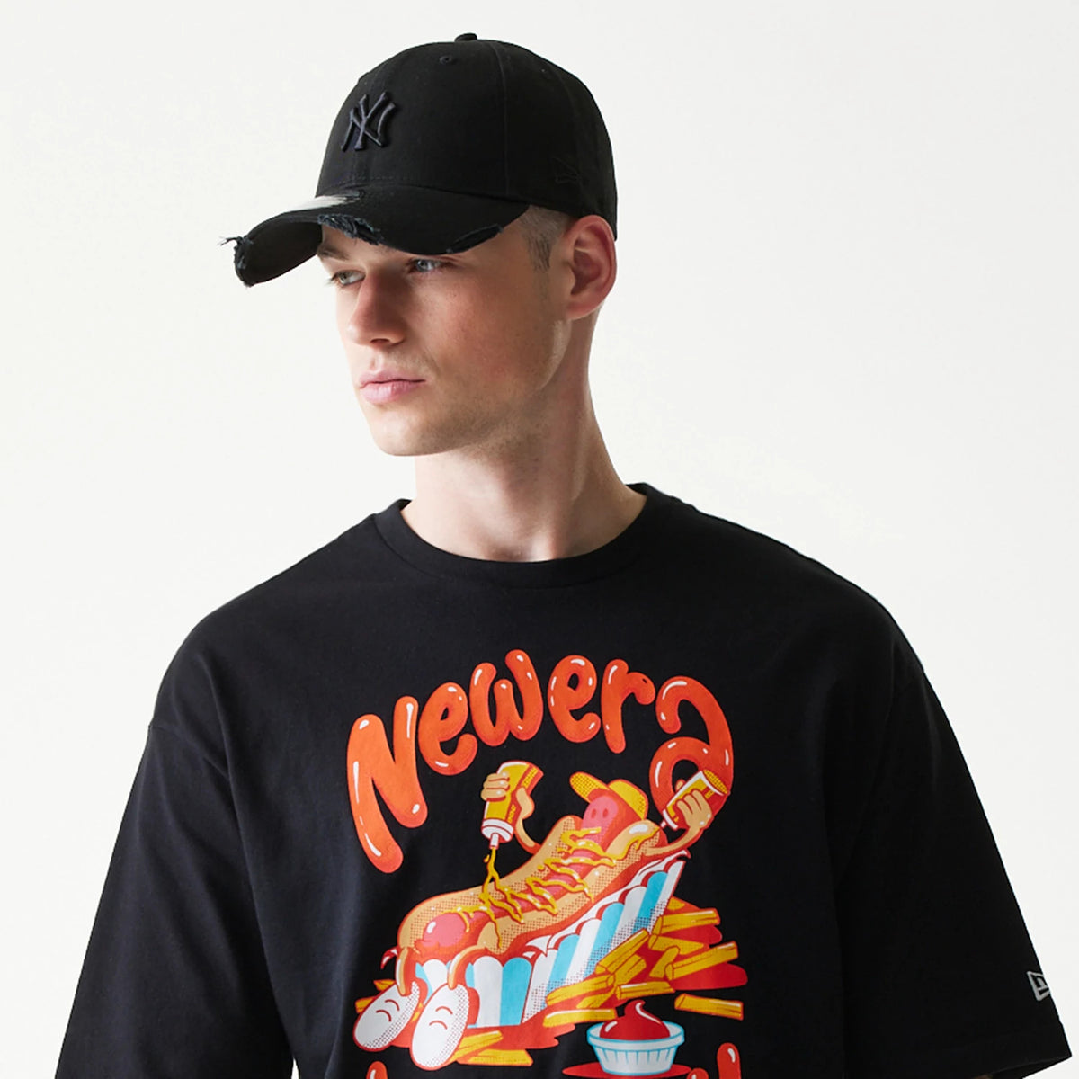 New Era Hotdog Food Graphic Black Oversized T-Shirt, front view on-model featuring a cartoon hotdog graphic with stacked chips and sauce, large red New Era lettering above, and a small embroidered logo on the left sleeve.