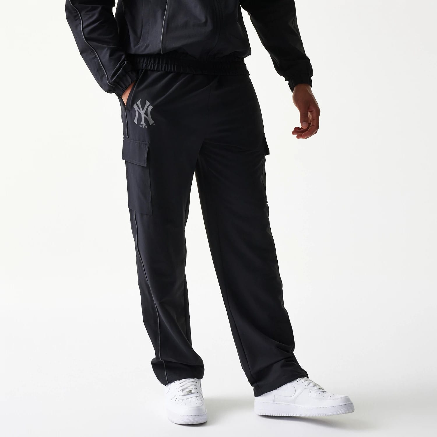 New Era New York Yankees MLB Utility Black Straight Leg Cargo Joggers, front view on a model wearing white trainers and a matching jacket, with the grey Yankees logo on the left thigh and cargo pockets on each side.