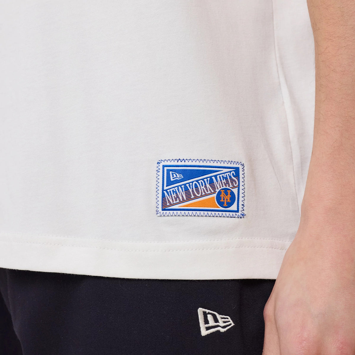 New Era New York Mets MLB Sport Classic White T-Shirt, side view showing an embroidered patch with New York Mets lettering and logo near the bottom hem, with a New Era logo on the model's black trousers.