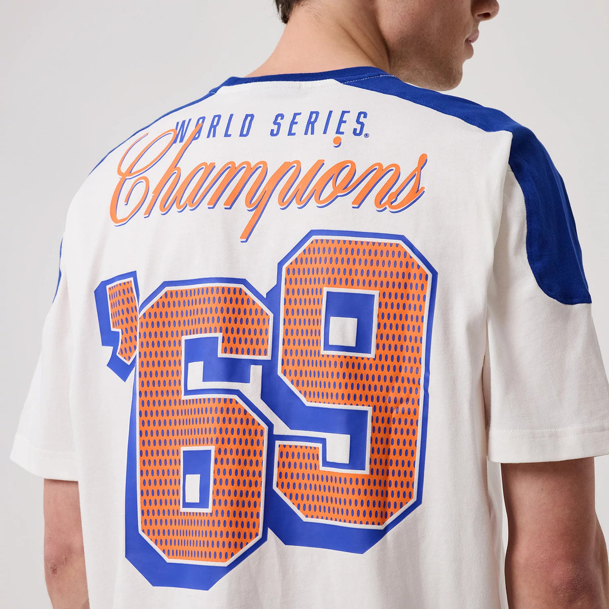 New Era New York Mets MLB Sport Classic White T-Shirt, back view on a model showing orange and blue '69' graphics beneath 'World Series Champions' lettering across the shoulders, with contrasting blue panels on the sleeves.