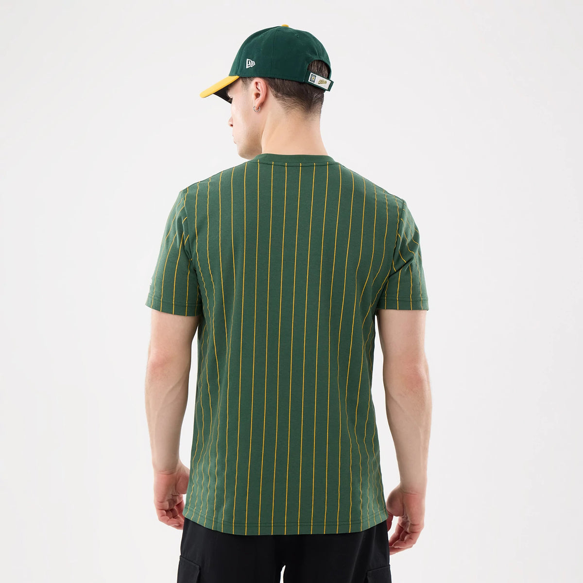 New Era Athletics MLB Pinstripe Dark Green T-Shirt, back view on a model showing vertical yellow pinstripes on a dark green background, styled with a matching green cap featuring an embroidered New Era logo on the side.