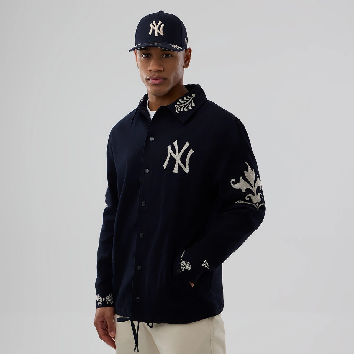 New Era New York Yankees MLB Script Navy Coach Jacket, front view on a model with a matching cap, showing an embroidered NY logo on the left chest, ornate designs on the collar and sleeves, and a small embroidered New Era logo on the wrist.