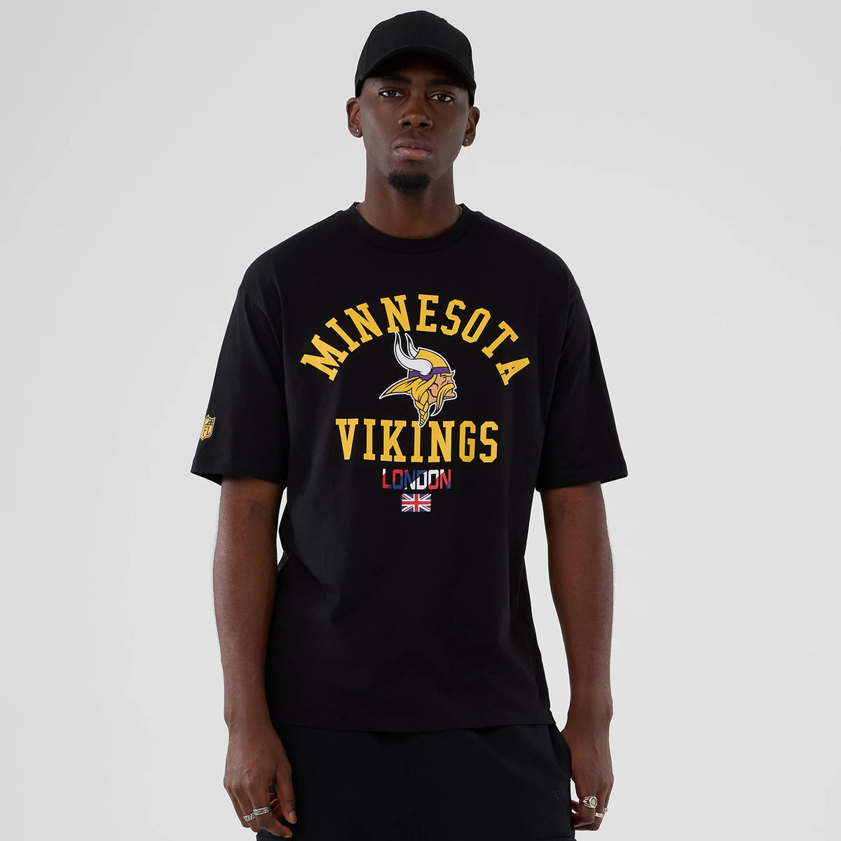 New Era Minnesota Vikings NFL International Series Games 2025 Black Oversized T-Shirt, front view on-model showing the yellow Vikings lettering and logo across the chest with a London motif and Union Jack below.