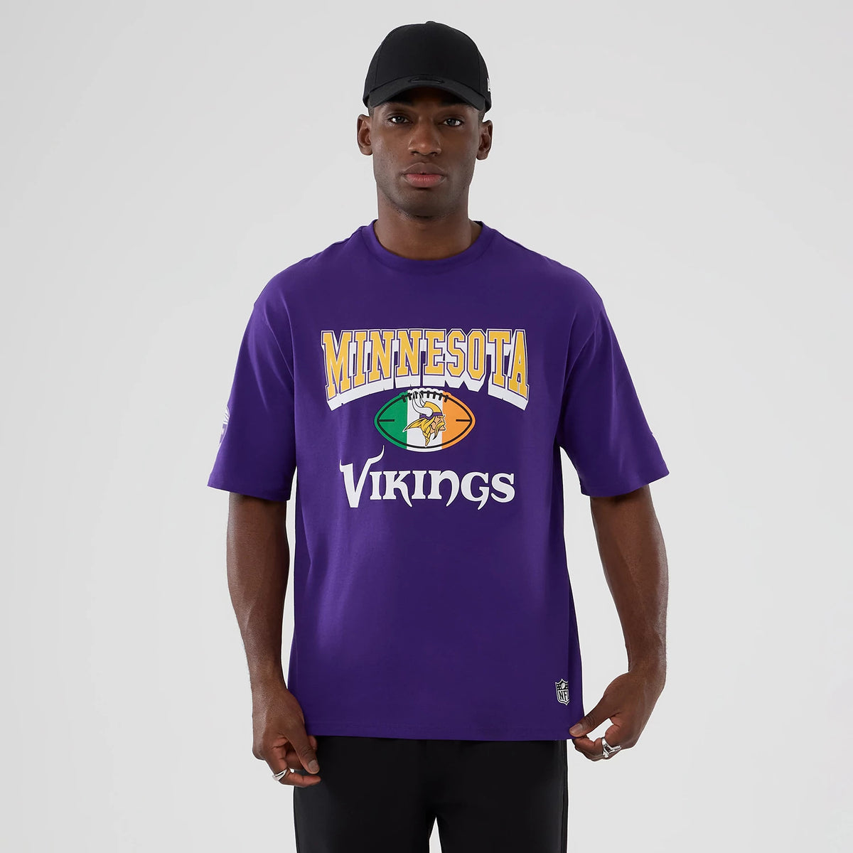 New Era Minnesota Vikings NFL International Series Games 2025 Purple Oversized T-Shirt, front view on-model displaying large printed Minnesota Vikings lettering in white and gold across the chest and a small embroidered NFL logo near the bottom hem.