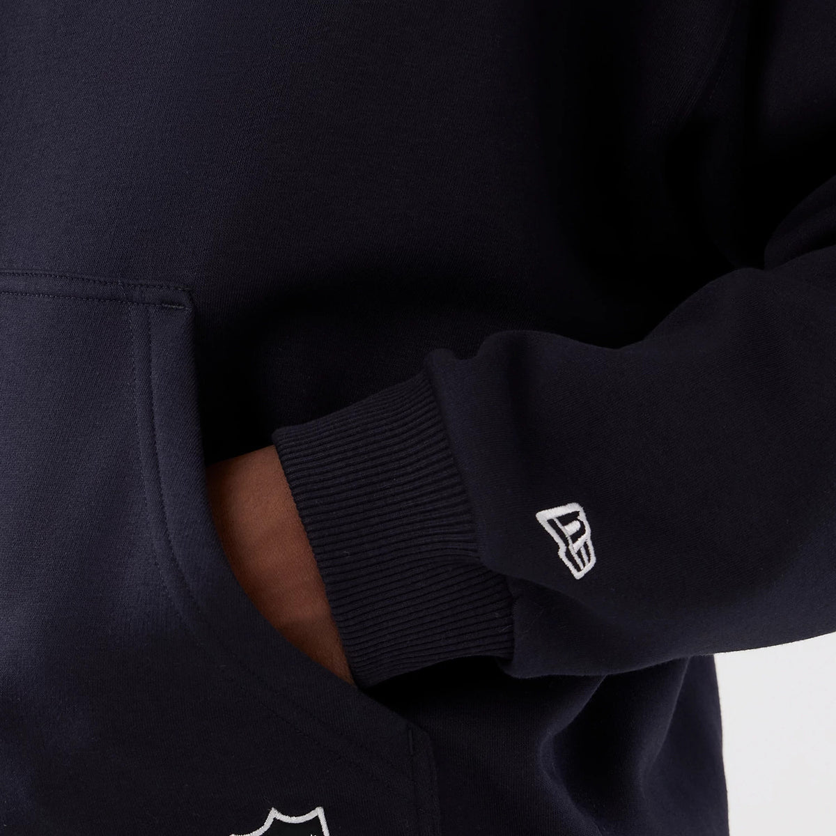 New Era Denver Broncos NFL International Series Games 2025 Navy Premium Pullover Hoodie, close-up view showing the pocket detail with an embroidered New Era logo on the sleeve and a portion of the NFL logo near the pocket.