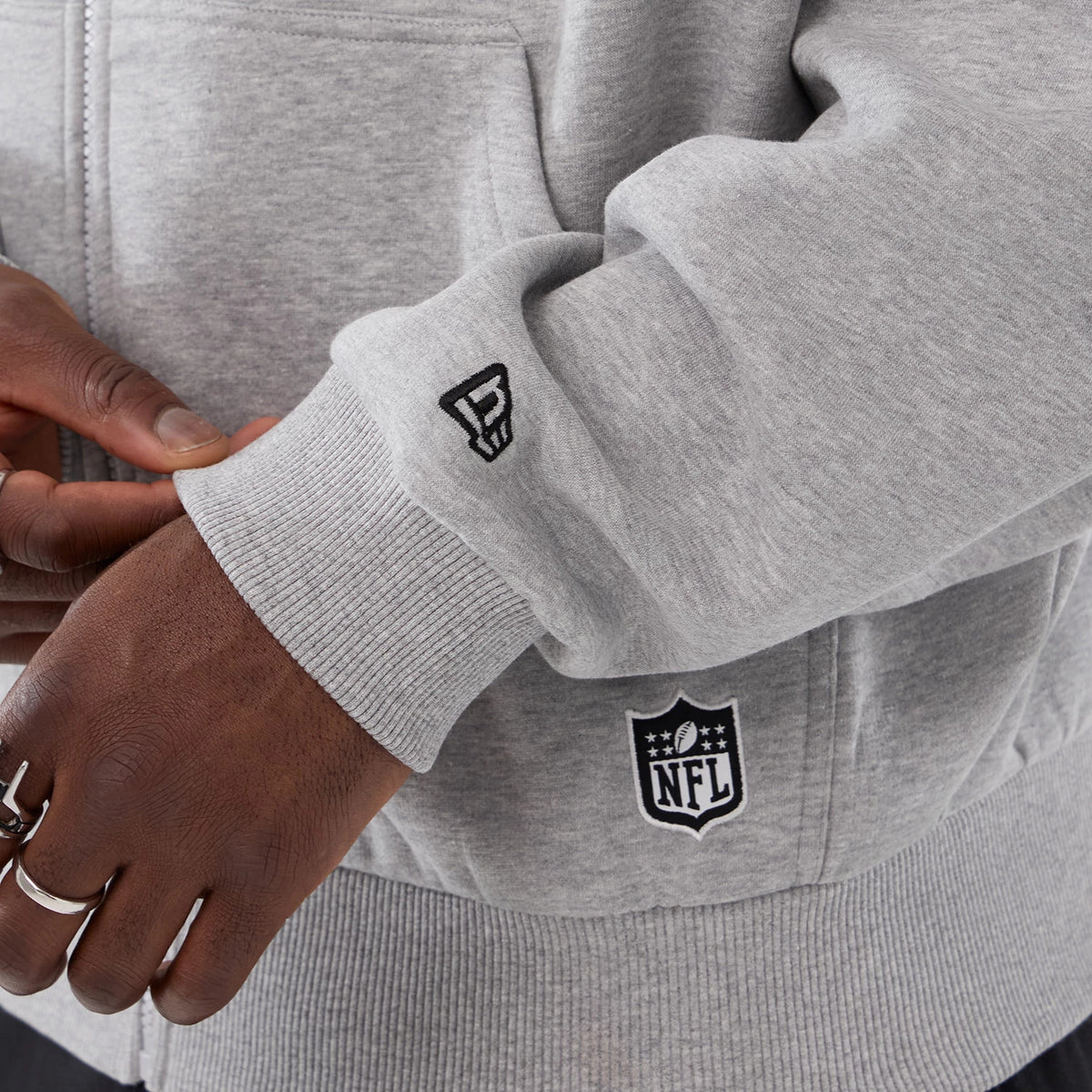 New Era Minnesota Vikings NFL International Series Games 2025 Grey Premium Pullover Hoodie, detail view showing a raised New Era logo on the sleeve cuff near the ribbed hem and an embroidered NFL logo on the bottom of the grey fabric.