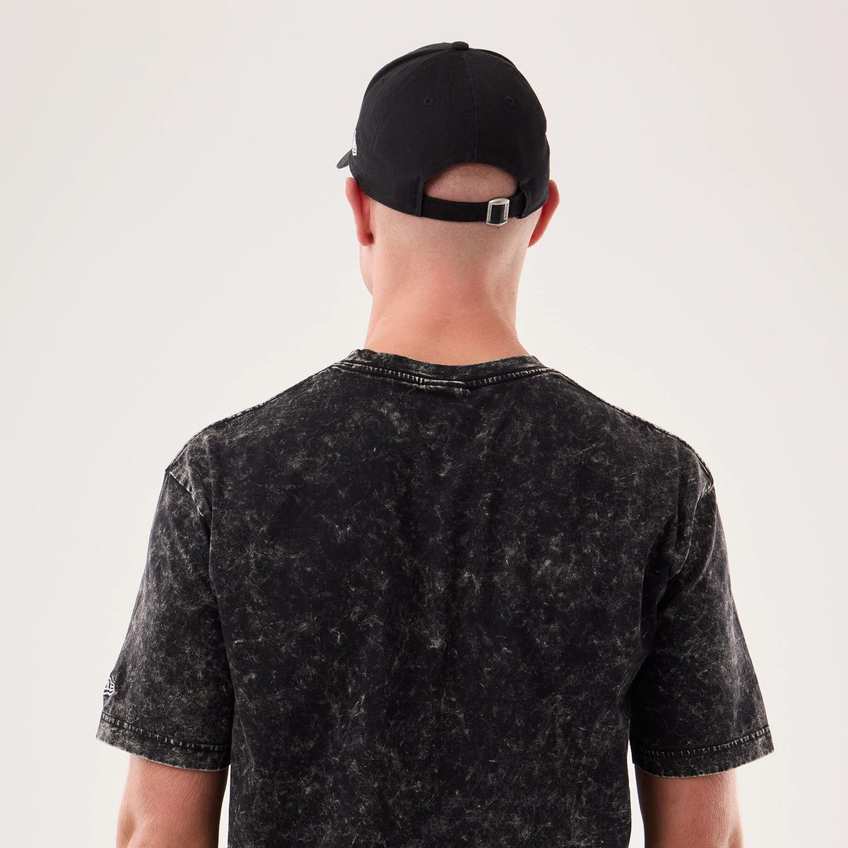 New Era Rick And Morty Science Black T-Shirt, back view on a model with a dark cap, shows a mottled black and grey pattern across the fabric, short sleeves, and an embroidered New Era logo on the left sleeve.