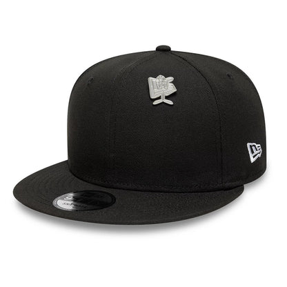 New Era Flag Character Silver Pin Badge, front view on a black cap showing a silver pin badge on the structured crown, an embroidered New Era logo on the side, and a curved black visor with a sticker on the top.