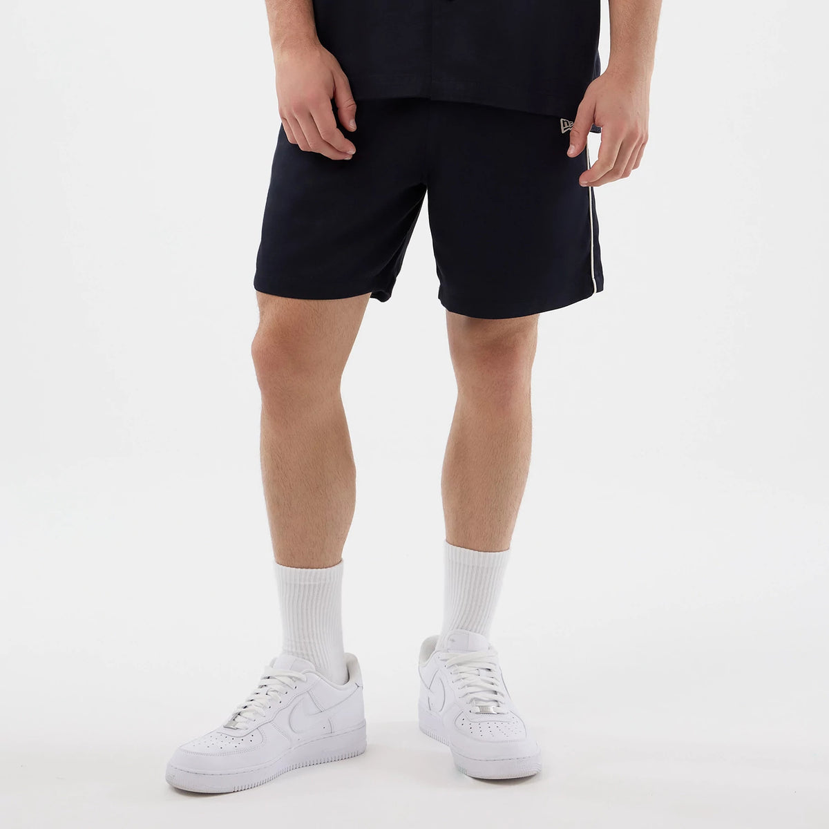 New Era Piping Navy Shorts, front view on-model featuring a straight fit that falls above the knee, white piping along the side seams, and a small embroidered New Era logo near the left hem, paired with white trainers and socks.