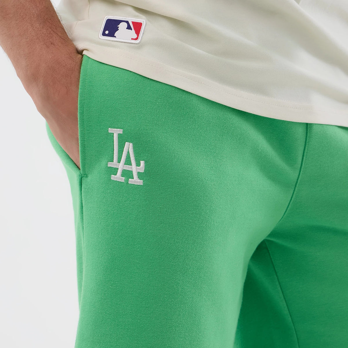 New Era LA Dodgers MLB League Essential Open Green Shorts, on-model view showing the vivid green shorts with a white embroidered LA logo on the left thigh, styled with a tucked-in cream shirt featuring a small MLB logo patch above the hem.