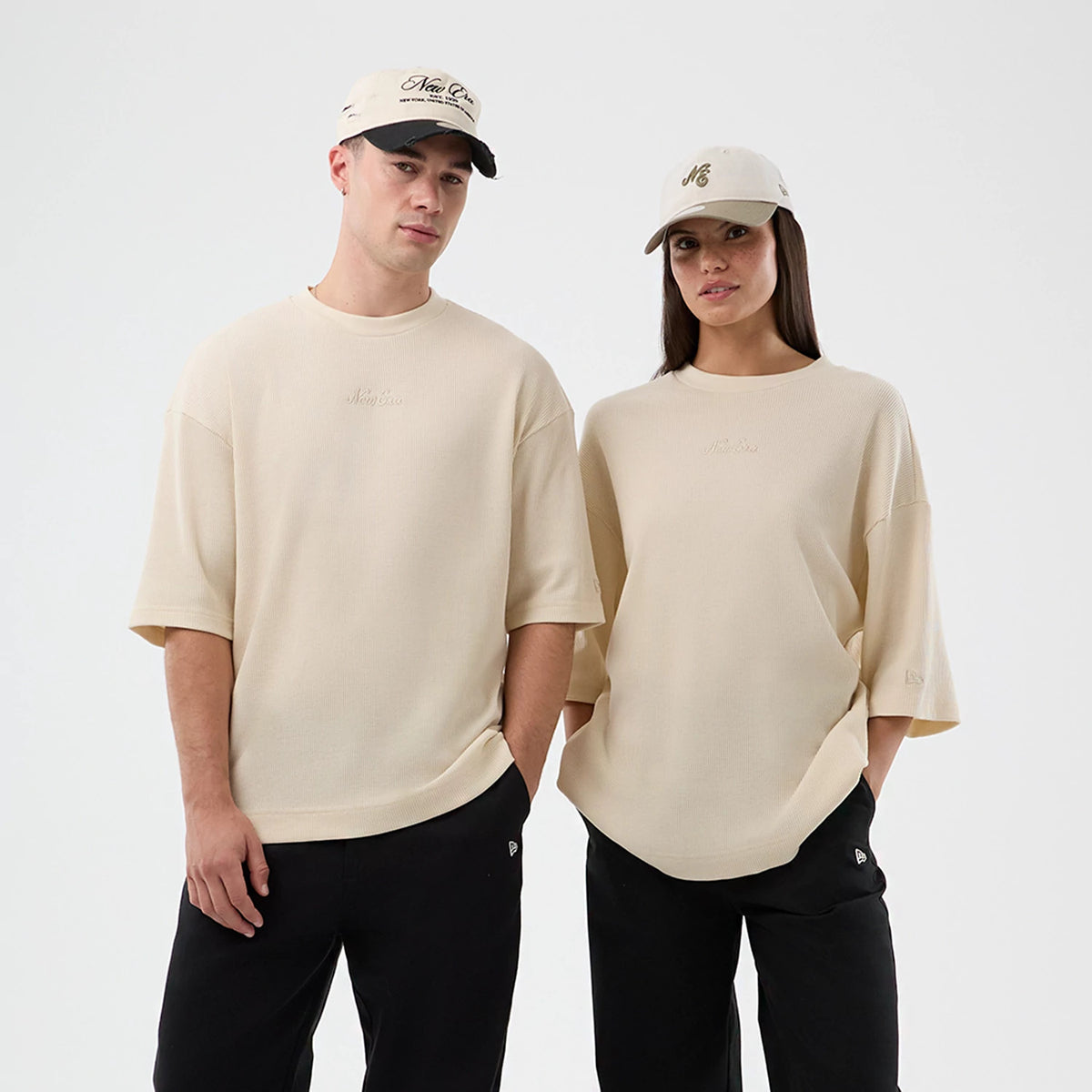 New Era Waffle Light Beige T-Shirt, front view on a model with a cap, showcasing the relaxed fit with mid-length sleeves, an embroidered New Era logo on the chest, and black trousers pairing the light beige tone.