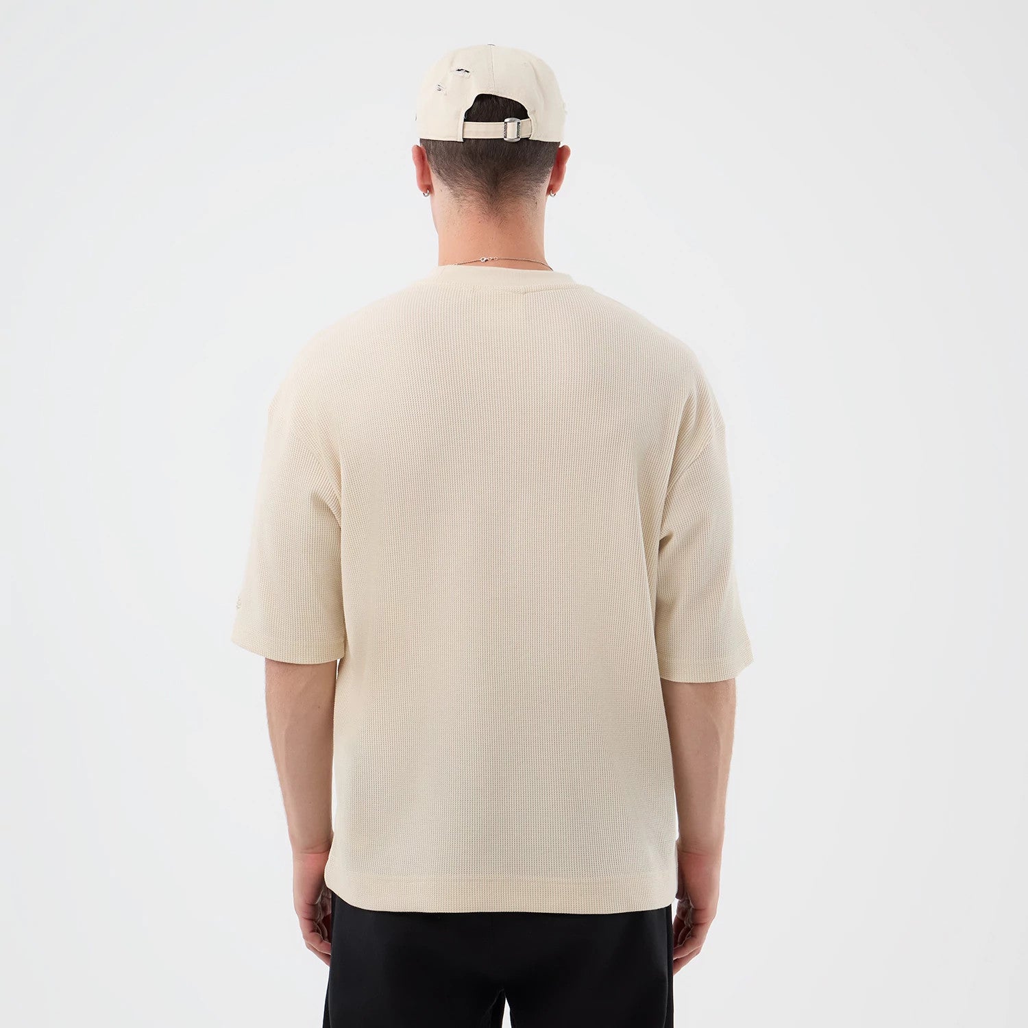 New Era Waffle Light Beige T-Shirt, back view on a model wearing a matching beige cap, showing the plain light beige textured fabric with short sleeves and a straight hem falling over the top of black trousers.