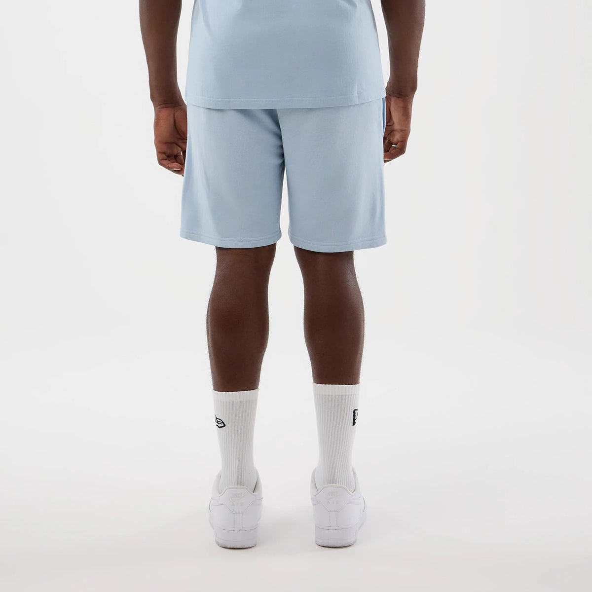 New Era LA Lakers NBA League Essential Pastel Blue Shorts, back view on a model showing the light blue shorts reaching above the knee, paired with a matching top, white socks with an embroidered New Era logo, and white trainers.