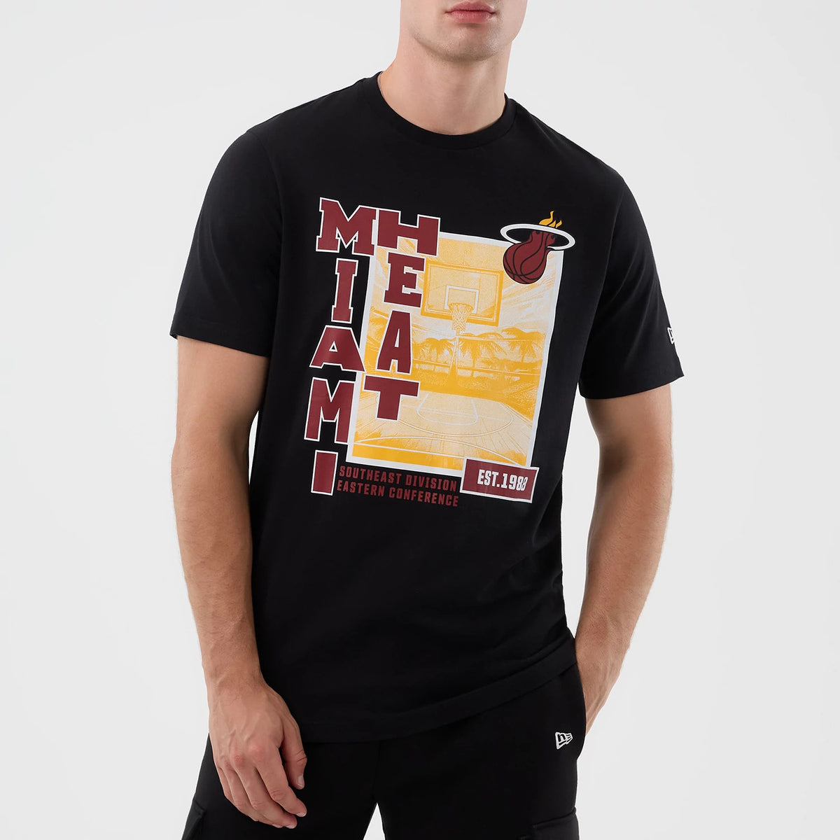 New Era Miami Heat NBA Graphic Black T-Shirt, front view on a model featuring large vertical MIAMI HEAT lettering in red on the left, a yellow and white graphic with a basketball court, and an embroidered New Era logo on the sleeve.