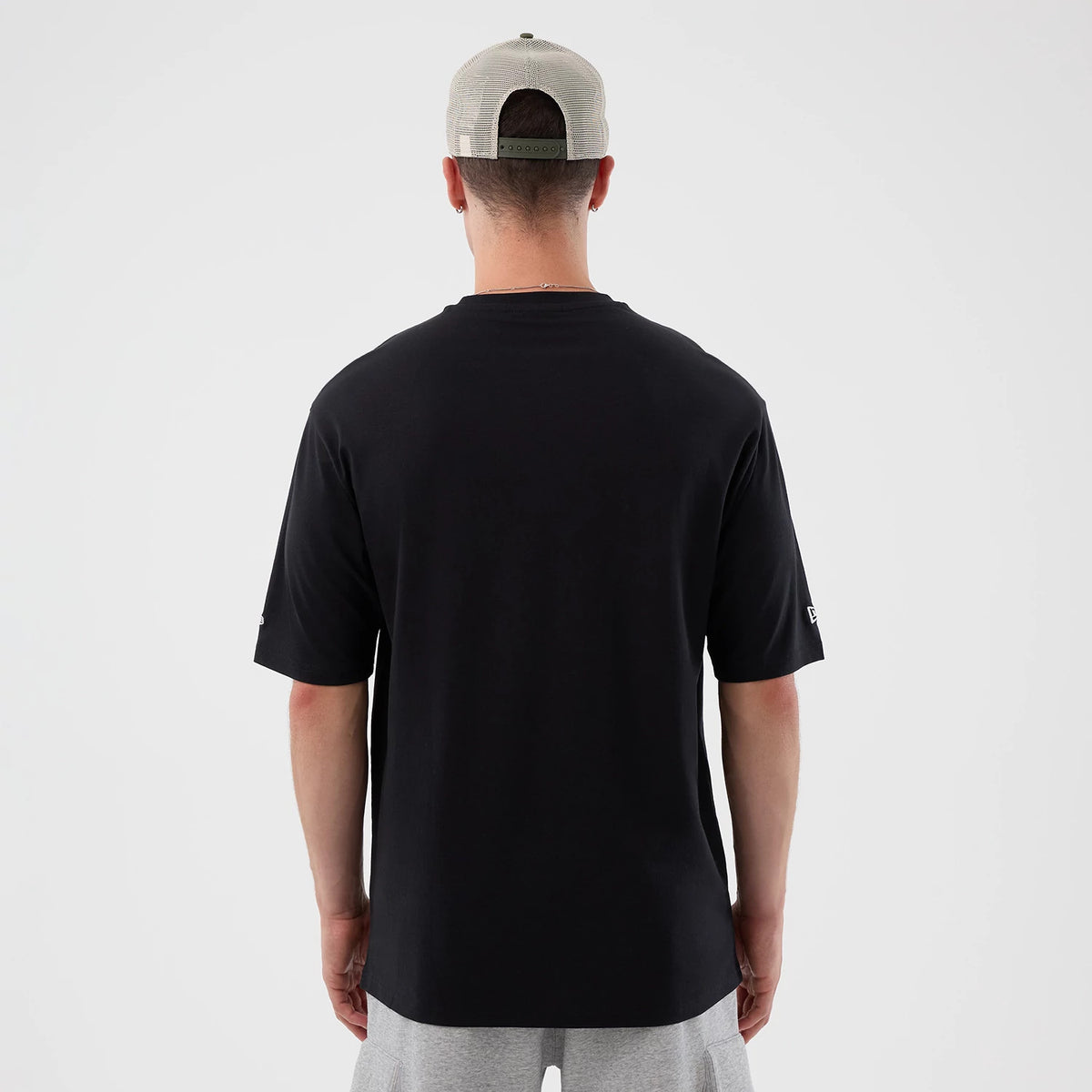 New Era New York Mets MLB Icon Graphic Black T-Shirt, back view on-model showcasing a plain black design with short sleeves, the New Era logo embroidered on the left sleeve, and the model styled with a beige cap and grey shorts.