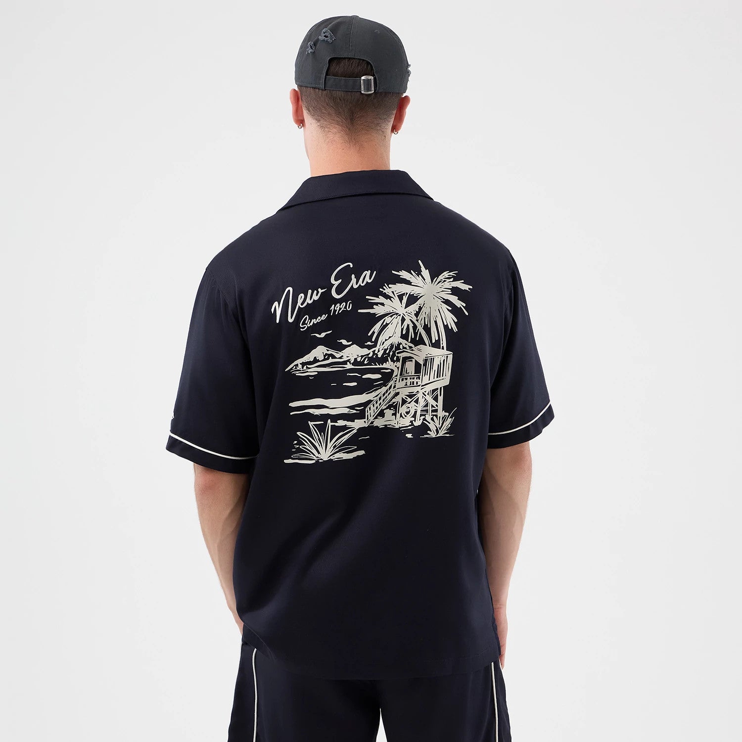 New Era Graphic Revere Navy Short Sleeve Shirt, back view on a model with a light design featuring a detailed beach scene, including palm trees and a cabin, and the text 'New Era Since 1920' in cursive script across the upper back.