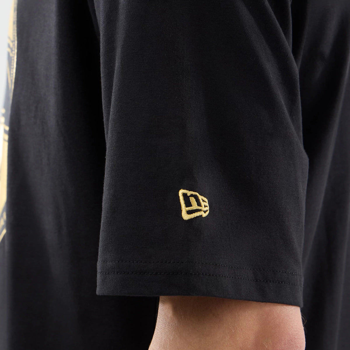 New Era Sticker Lisbon Black Oversized T-Shirt, on-model view focusing on the short sleeve, featuring a prominent yellow embroidered New Era logo near the hem and a glimpse of a light graphic on the body of the shirt.