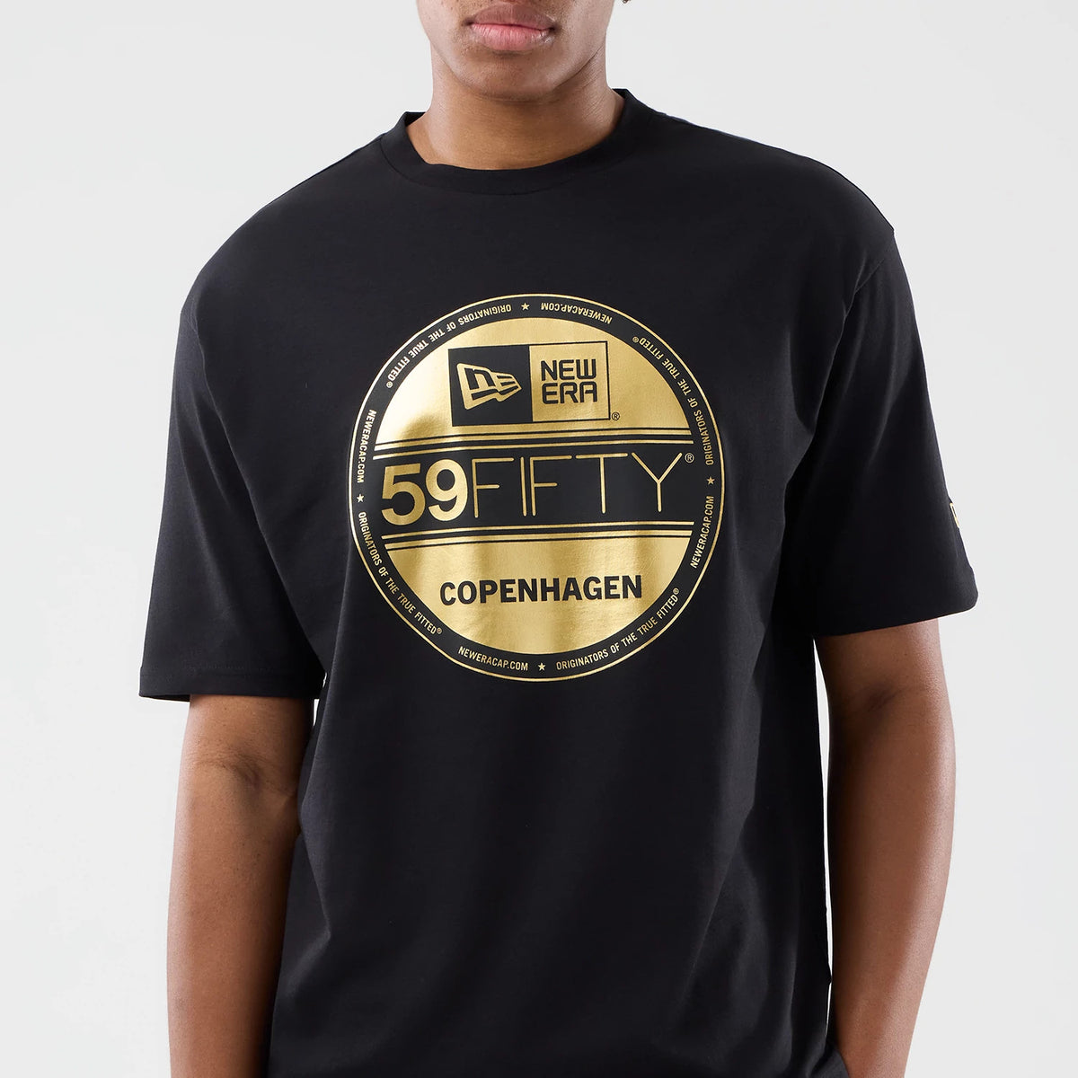 New Era Sticker Copenhagen Black Oversized T-Shirt, front view featuring a large gold-printed circular sticker design in the centre with New Era and 59FIFTY logos, Copenhagen text below, and short sleeves styled on a model.