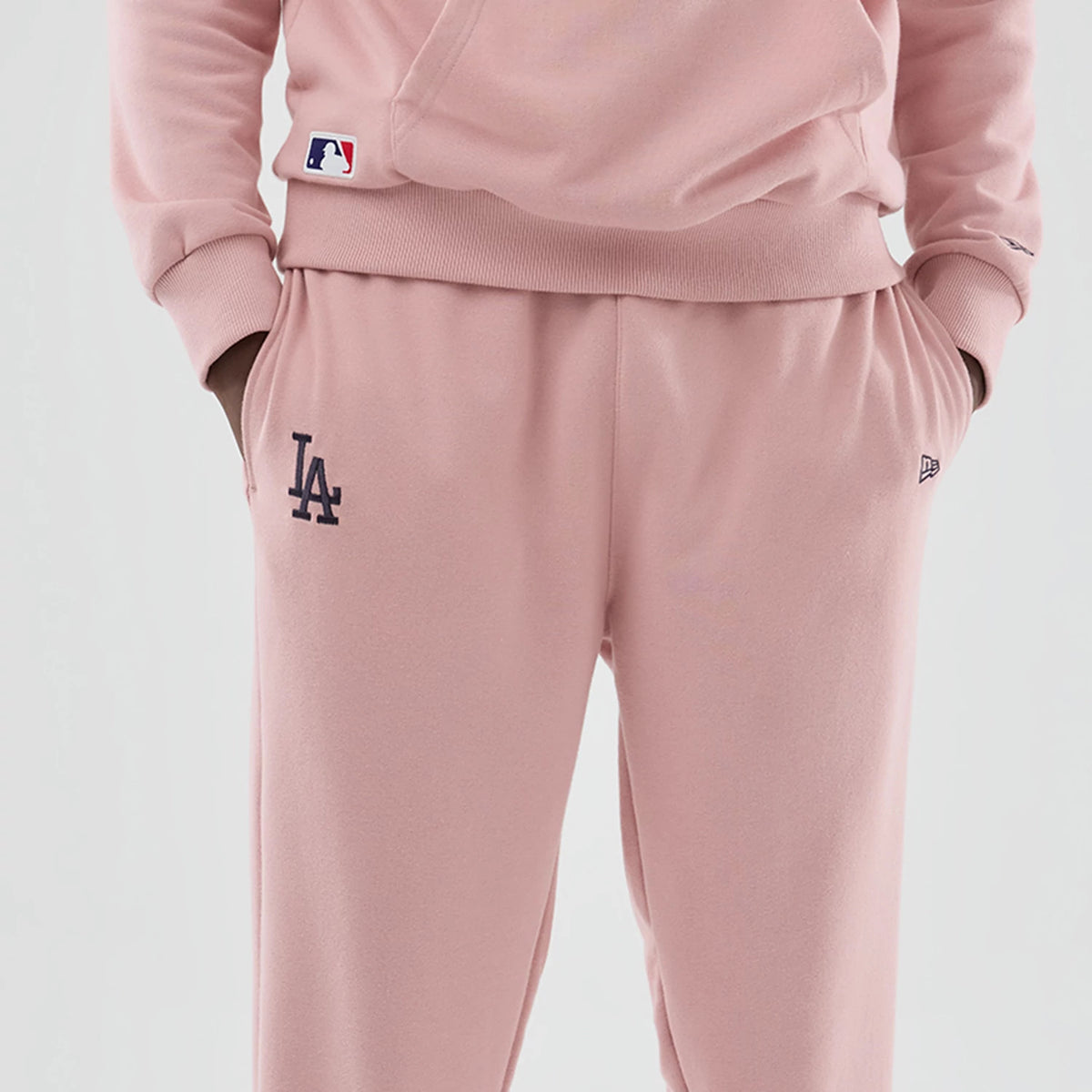 New Era LA Dodgers MLB Midi League Essential Pastel Pink Joggers, front view on a model showing the navy embroidered LA logo on the left leg, elasticated waistband for comfort, and the New Era flag logo on the right pocket.