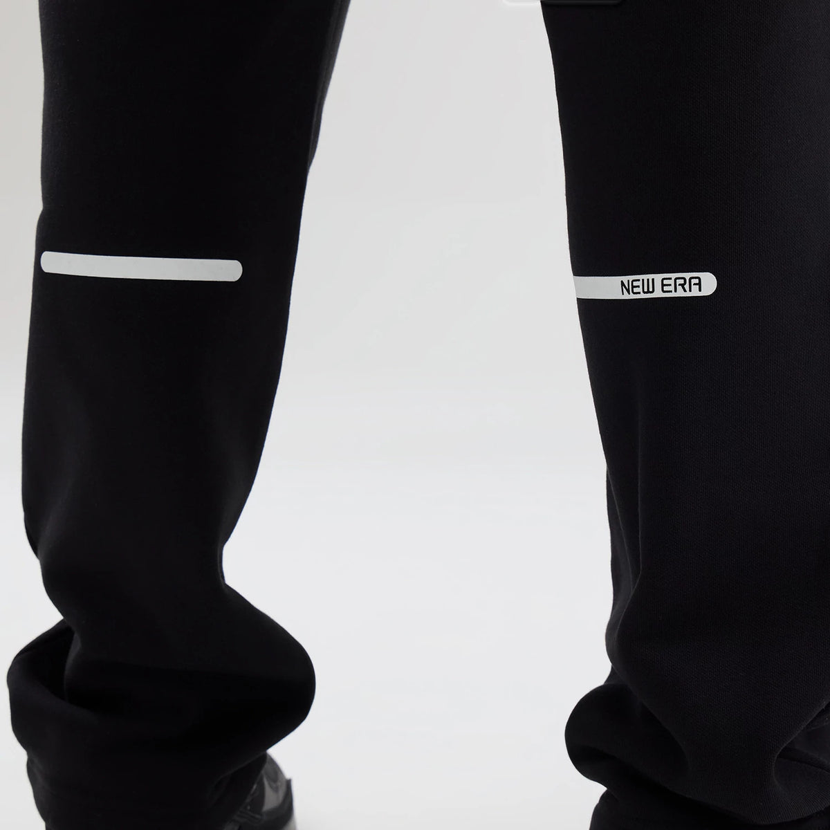 New Era Tech Reflective Black Straight Leg Joggers, close-up view showing reflective strips above the knee, with a white New Era logo on the right leg and black fabric for a modern look.