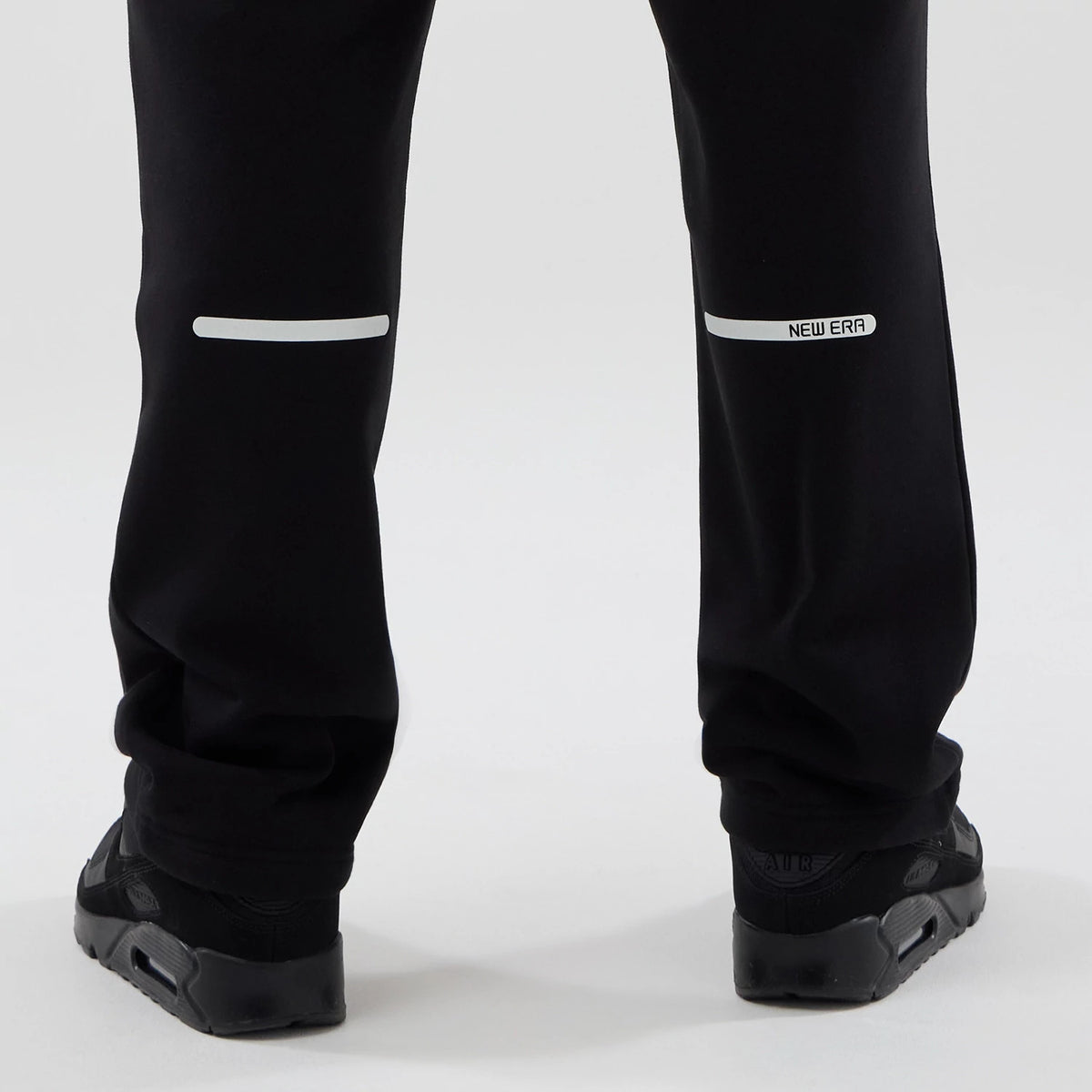 New Era Tech Reflective Black Straight Leg Joggers, rear view showing reflective strips across the lower legs and a New Era logo near the right strip, with a relaxed fit over black trainers.
