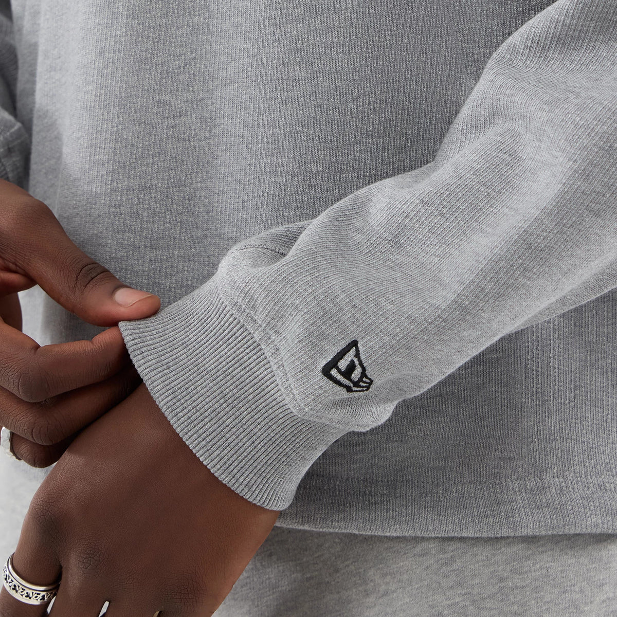 New Era Grey Oversized Crew Neck Sweater, close-up view showing a ribbed cuff with a small black embroidered New Era flag logo, grey fabric texture with knit pattern, and the sweater’s relaxed fit design.