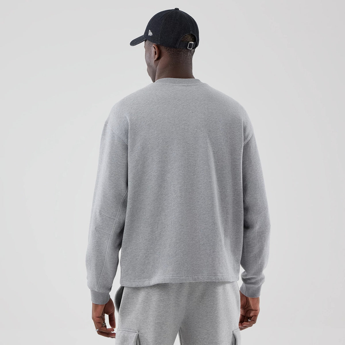 New Era Grey Oversized Crew Neck Sweater, back view on a model wearing matching grey sweatpants and a black cap with the New Era flag logo on the left side, highlighting the sweater’s simple design and relaxed fit.