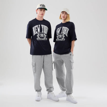 New Era New York Yankees MLB Arch Graphic Navy Oversized T-Shirt, front view on-model showing large white New York lettering with the Yankees logo in the centre, complemented by grey cargo trousers and white trainers, with caps adding a casual style.