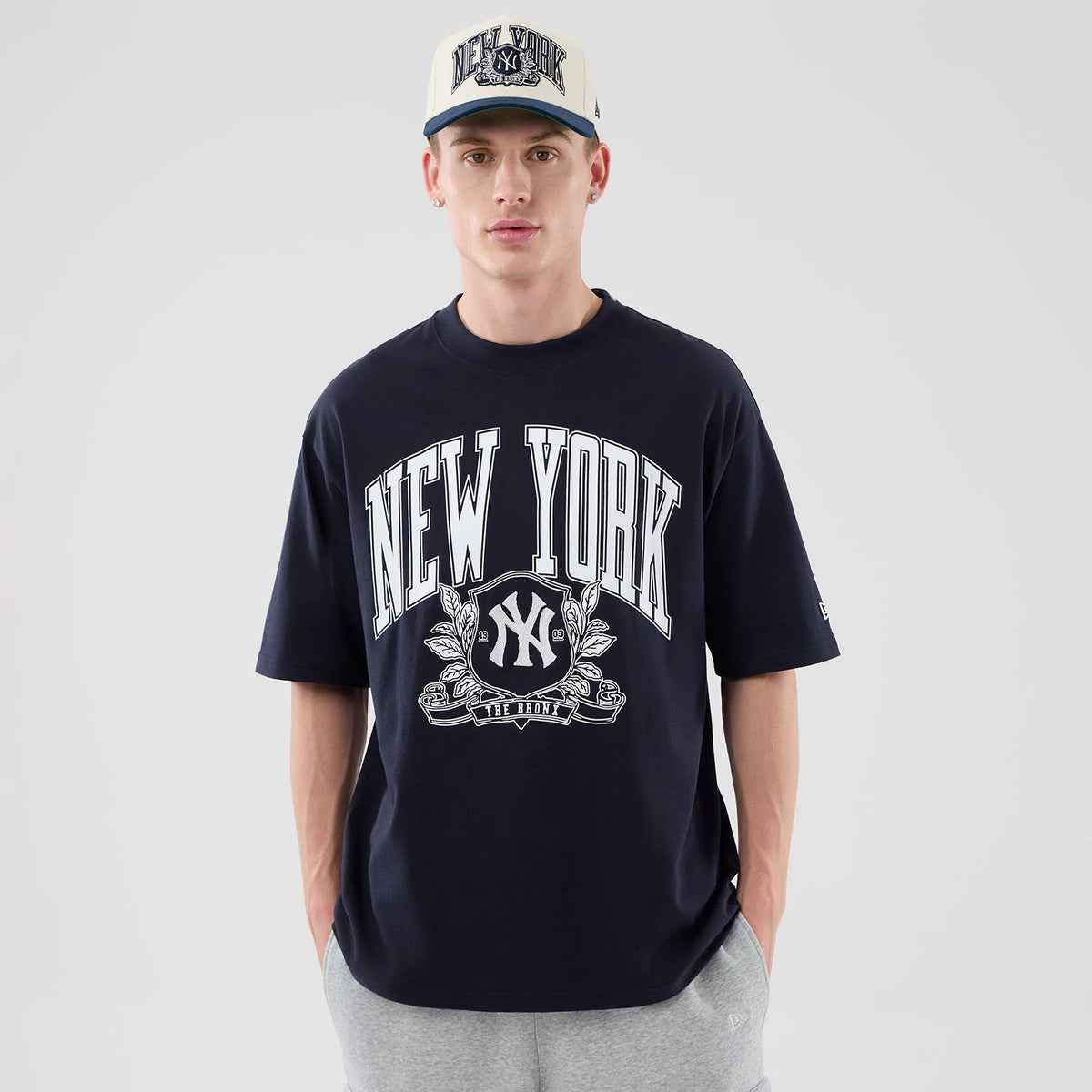 New Era New York Yankees MLB Arch Graphic Navy Oversized T-Shirt, front view on a model with a white cap, displaying a large New York Yankees graphic across the chest, with white lettering and the embroidered logo centred below it.