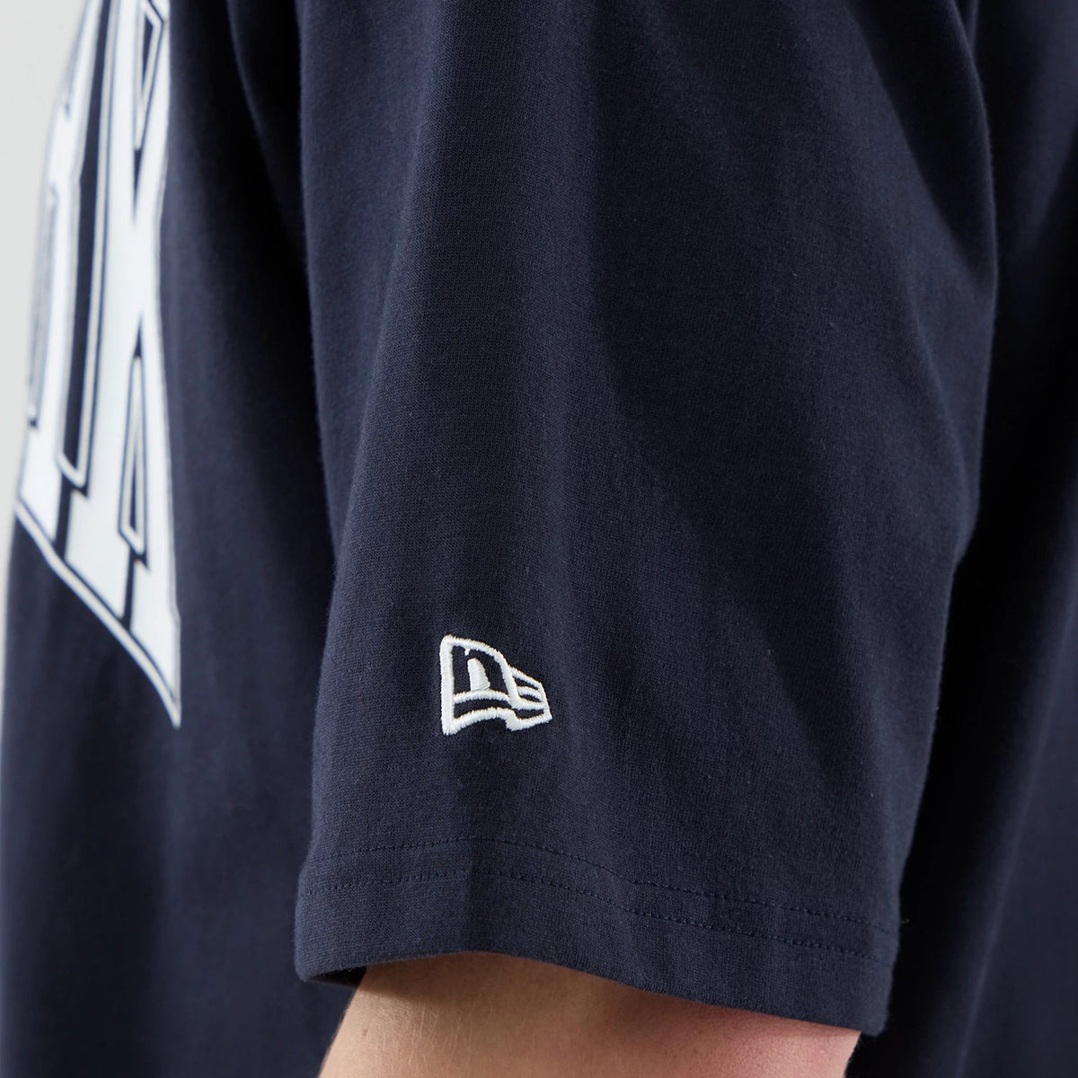 New Era New York Yankees MLB Arch Graphic Navy Oversized T-Shirt, side view showing the sleeve with a white embroidered New Era logo near the hem, and part of the oversized white lettering graphic across the back, set against the navy fabric.