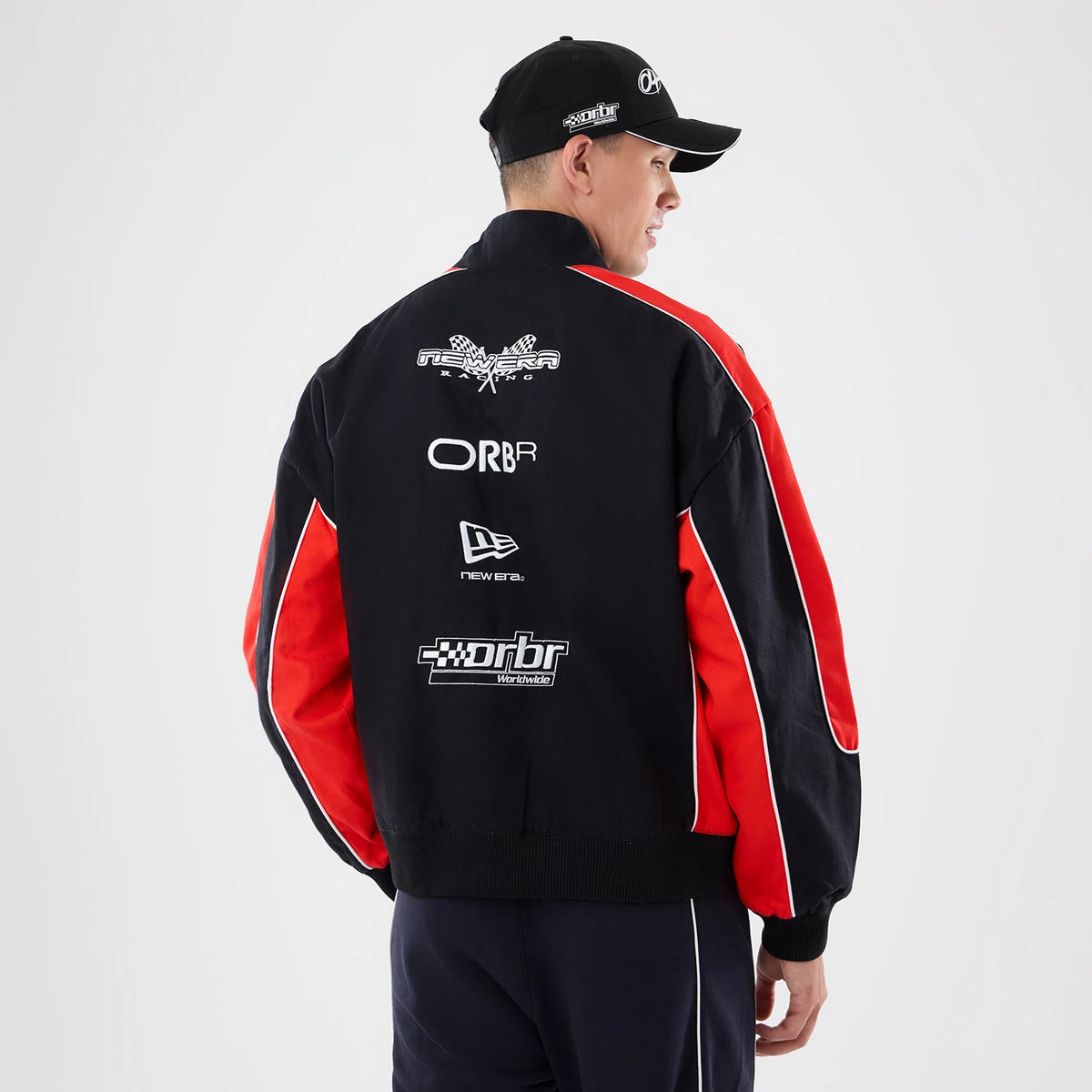 New Era Red Bull Racing Motorsport Black Jacket, back view on a model wearing a black cap, featuring multiple white embroidered logos including New Era, ORBR, and Red Bull Racing on the upper back, and red and white striped on the sleeves.