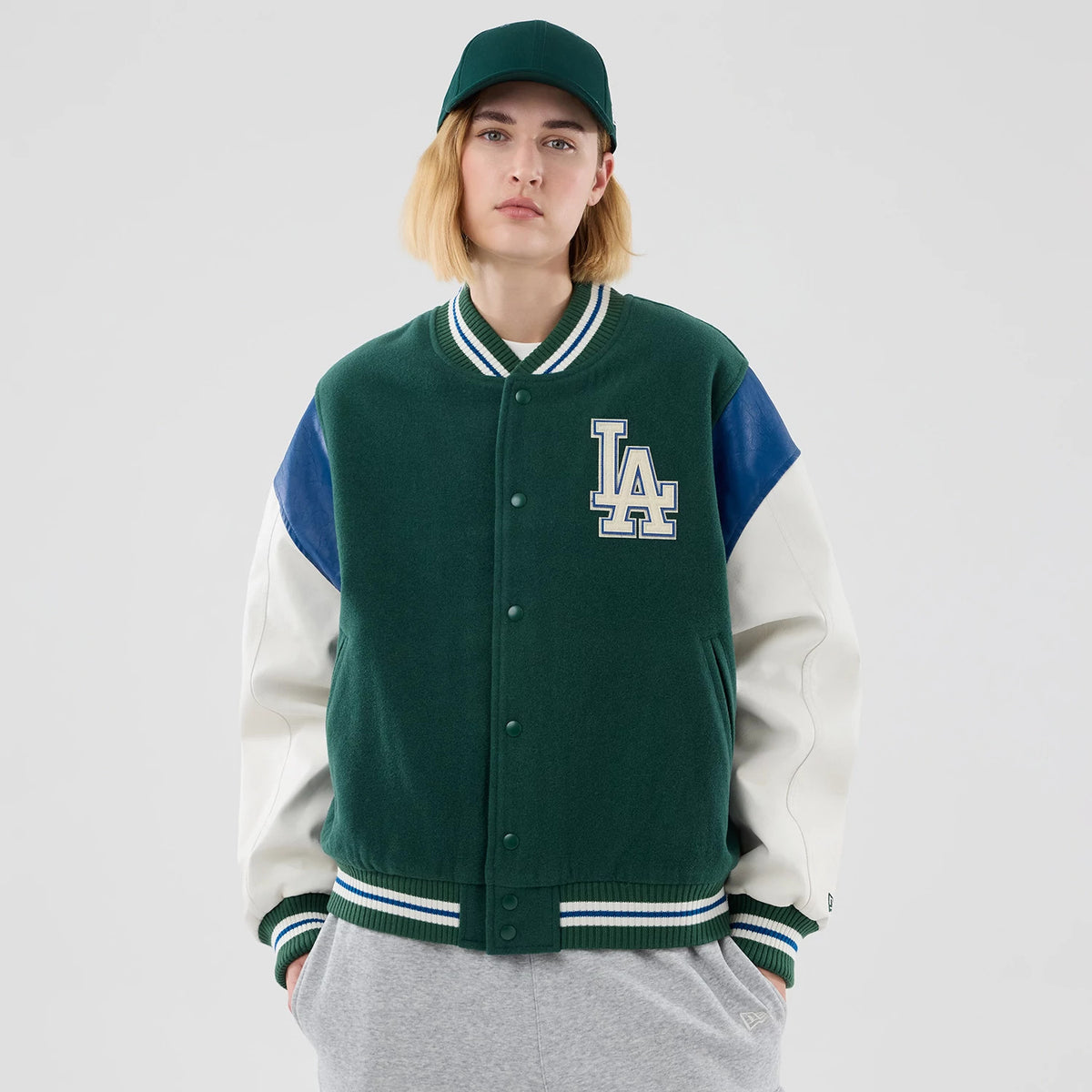 New Era LA Dodgers MLB Varsity Dark Green Jacket, front view on a model featuring a large embroidered LA logo on the left chest, contrasted white sleeves with blue accents, and ribbed green and white striped cuffs and collar.