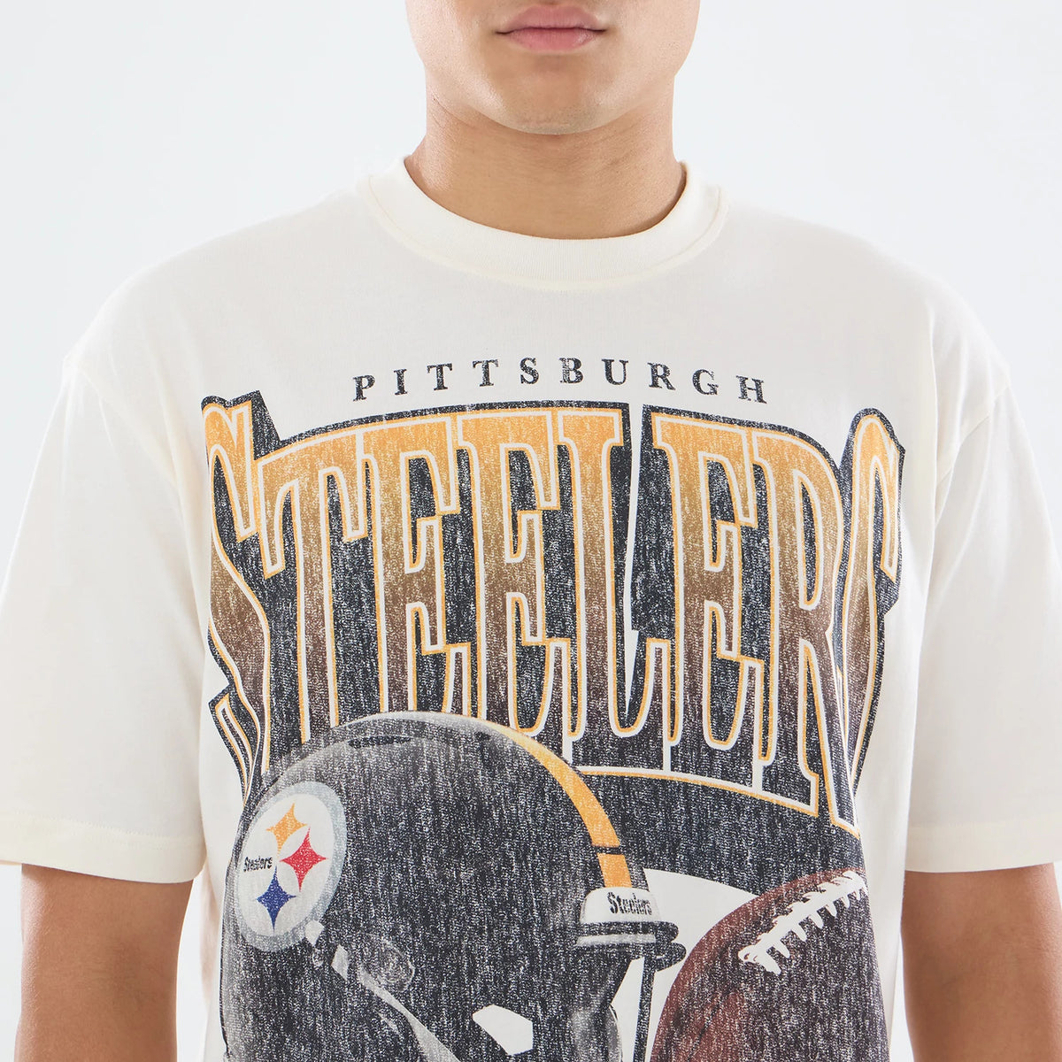 New Era Pittsburgh Steelers NFL Sport Classic Cream T-Shirt, front view on a model showing a printed design with large Steelers lettering across the chest, a detailed print of a football helmet with team logo beneath, and a faded vintage effect.