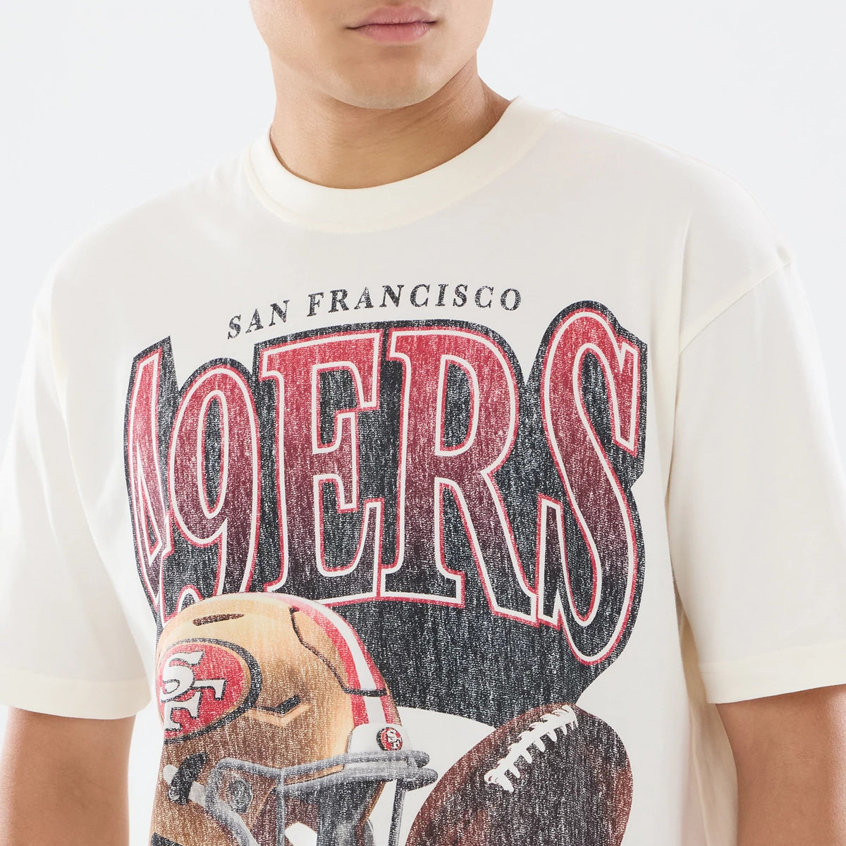 New Era San Francisco 49ers NFL Sport Classic Cream T-Shirt, front view on a model featuring large red and black 49ERS lettering across the chest, SAN FRANCISCO text above, and a detailed graphic of a 49ers helmet and football below the lettering.