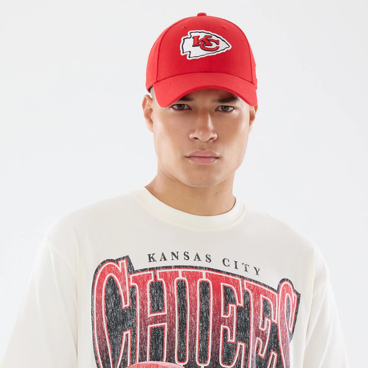 New Era Kansas City Chiefs NFL Sport Classic Cream T-Shirt, front view on a model wearing a red cap, featuring CHIEFS lettering printed across the chest in distressed red and black, and KANSAS CITY text above in smaller black font.