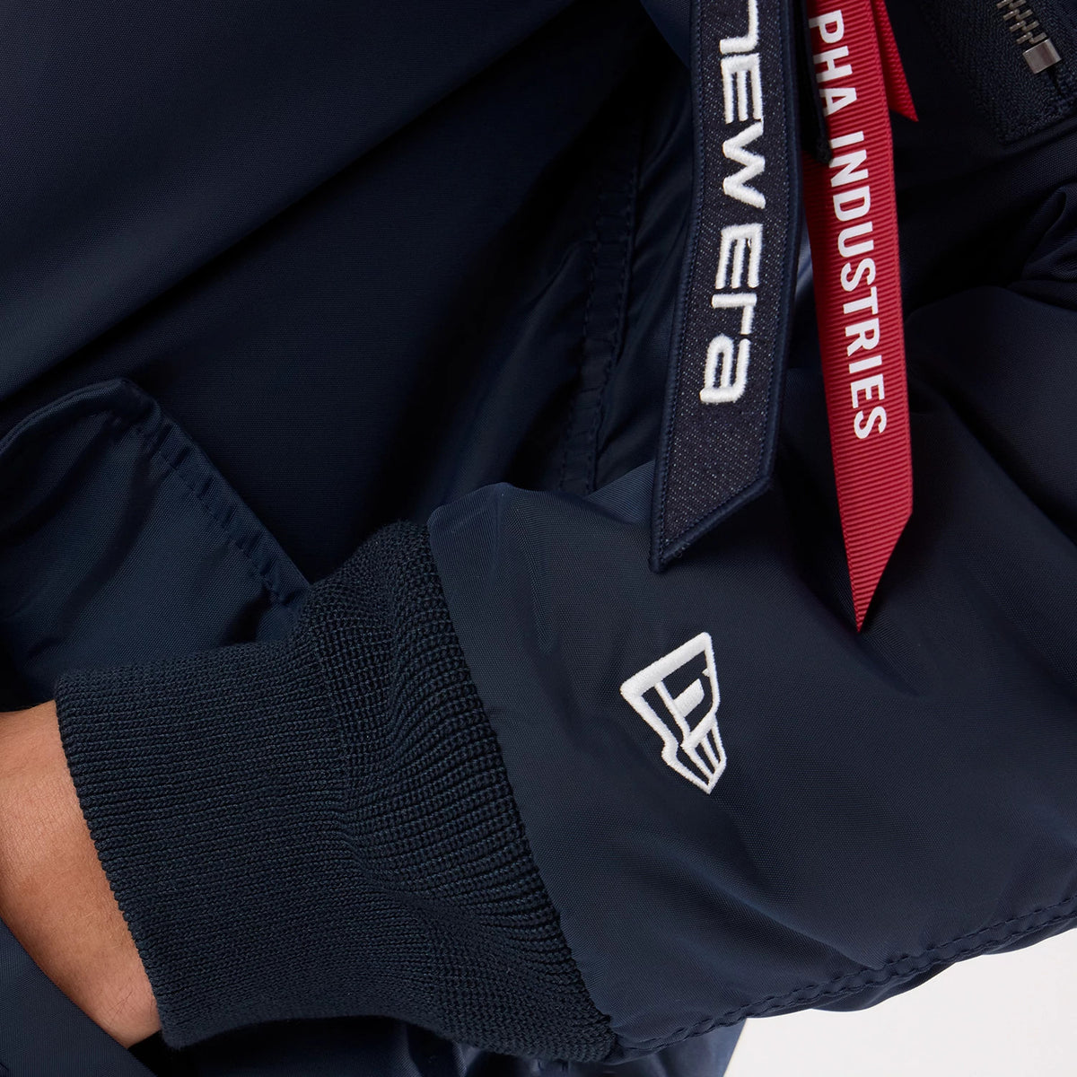 New Era New York Yankees MLB X Alpha Navy Jacket, close-up side view showing a white embroidered New Era logo near the cuff, a dark ribbed sleeve end, and red Alpha Industries tag detail with contrasting white lettering hanging from the zip.
