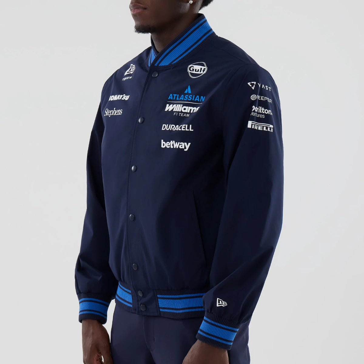 New Era Williams Navy Varsity Jacket, three-quarter view showing embroidered sponsor logos on the left chest and sleeves, a button-up front with blue accents on the striped collar, cuffs, and hem, and a white New Era logo on the left cuff.