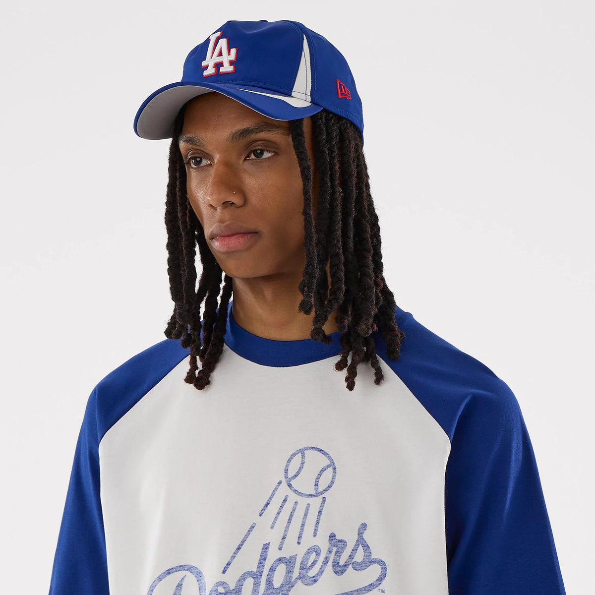 New Era LA Dodgers MLB White Boxy T-Shirt, on-model front view highlighting the large Dodgers script with a decorative baseball graphic on the chest, blue raglan sleeves, and styled with a two-tone blue and white cap featuring an embroidered LA logo.
