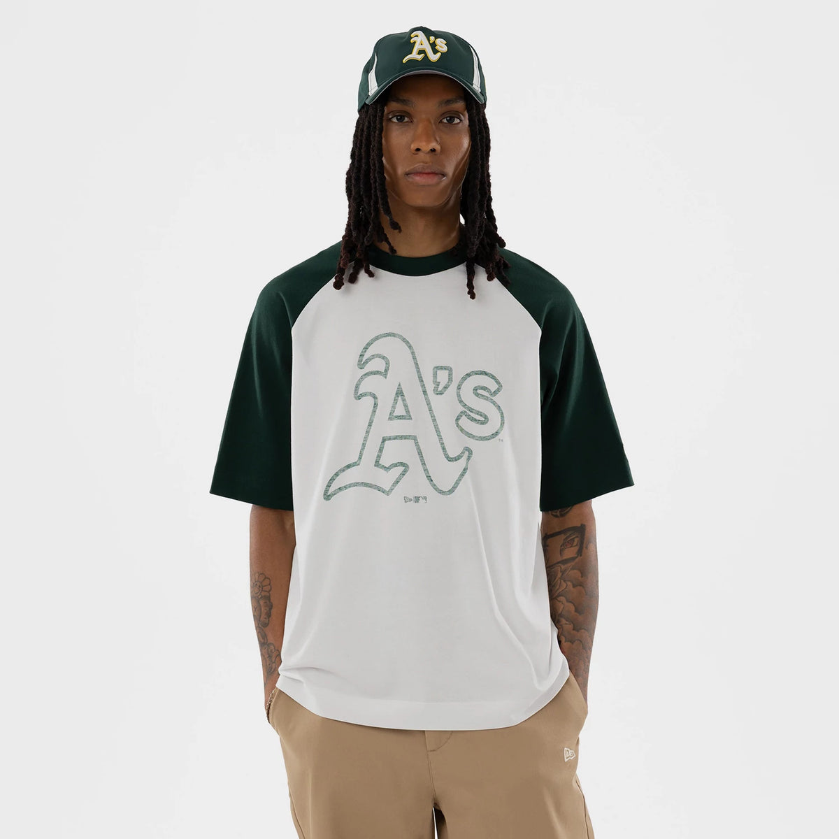New Era Athletics MLB White Boxy T-Shirt, front view showing a large embroidered ‘A’s’ logo in green on the centre, with contrasting dark green sleeves, worn by a model with beige trousers and a matching cap featuring an embroidered ‘A’s’ logo.