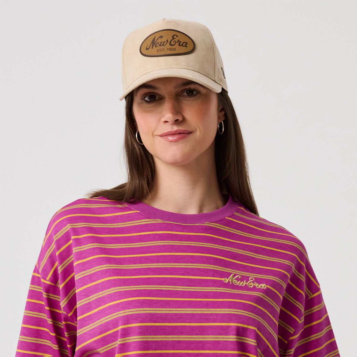 New Era Stripe Bright Purple T-Shirt, front view on-model showing bright purple and mustard stripes across the fabric, a yellow embroidered New Era logo on the left chest, and a complementary beige cap featuring a brown embroidered patch.