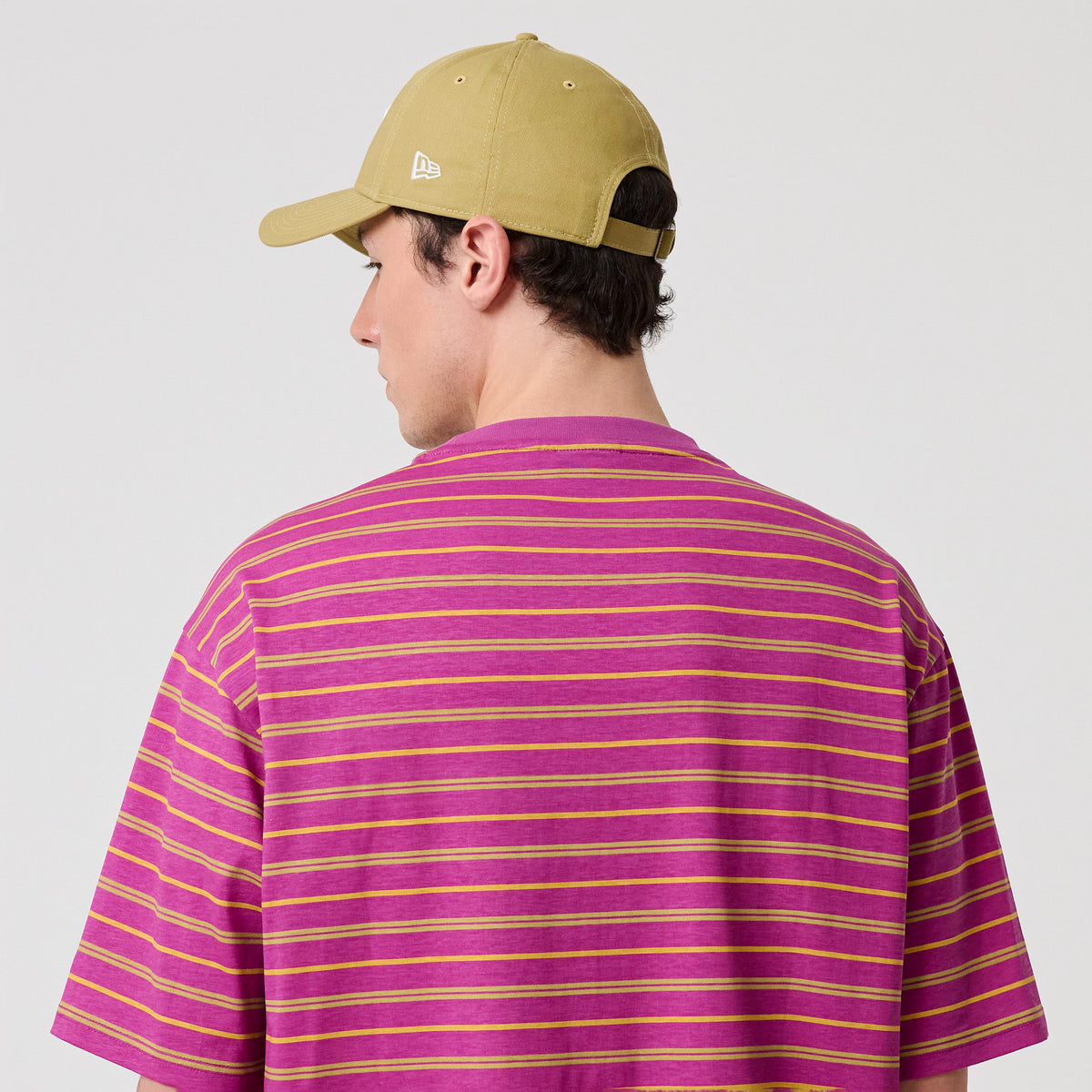 New Era Stripe Bright Purple T-Shirt, back view showing alternating yellow and pink horizontal stripes across the fabric, worn on-model with a beige cap featuring an embroidered New Era logo at the side.