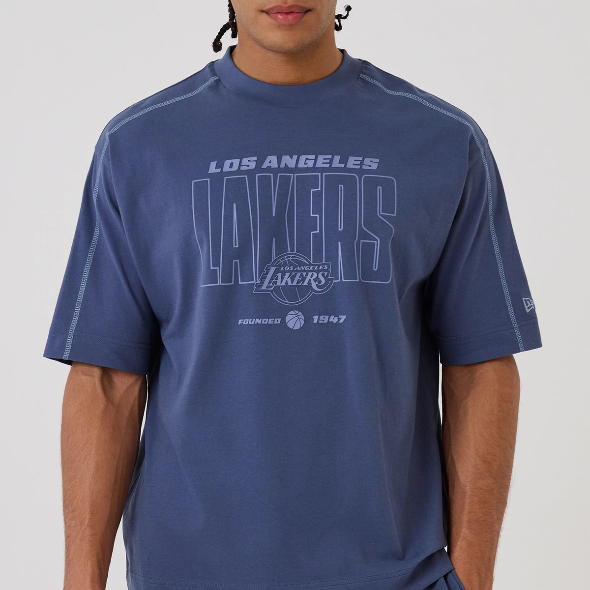 New Era LA Lakers NBA Graphic Dark Blue Boxy T-Shirt, front view on a model featuring large Los Angeles Lakers lettering across the chest, the embroidered New Era logo on the left sleeve, and contrast stitching along the shoulder seams.