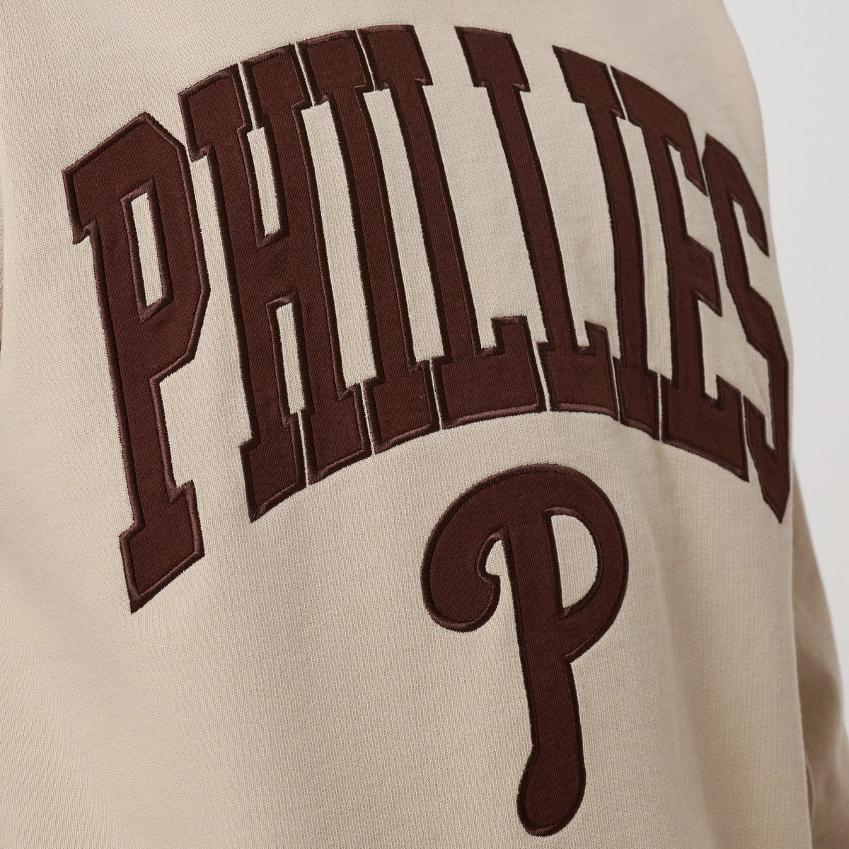 New Era Philadelphia Phillies MLB Arch Graphic Light Beige Crew Neck Sweater, close-up view showing large brown embroidered PHILLIES lettering in an arch design across the chest, with a prominent brown P below on light beige fabric.