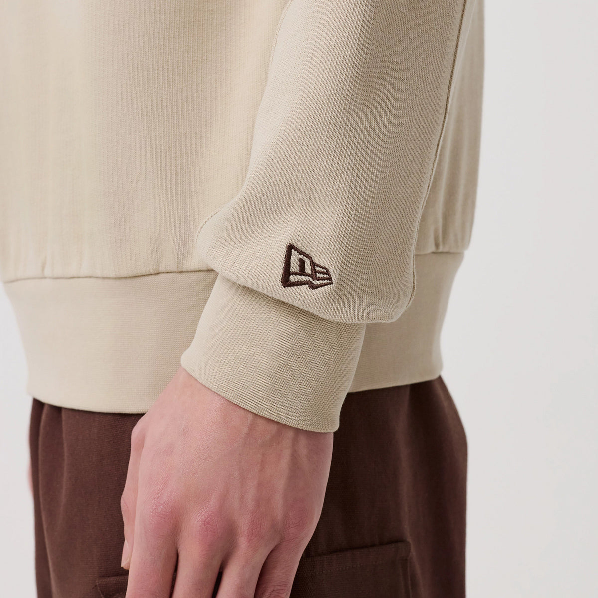New Era Philadelphia Phillies MLB Arch Graphic Light Beige Crew Neck Sweater, side view on a model showing the ribbed cuff with a brown embroidered New Era flag logo on the sleeve, paired with brown trousers for a casual look.