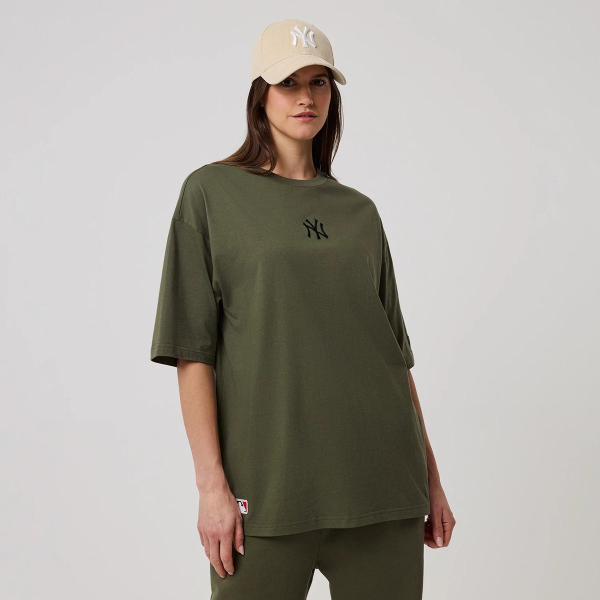 New Era New York Yankees MLB League Essential Green Oversized T-Shirt, front view on a model pairing it with a beige cap, featuring the black embroidered New York Yankees logo centred on the chest and a small branded label on the lower hem.