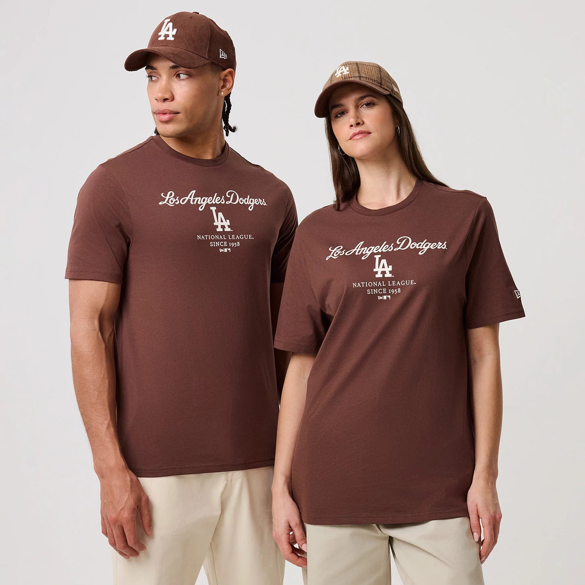 New Era LA Dodgers MLB Linear Brown T-Shirt, front view on-model showing the brown T-shirt with white printed Los Angeles Dodgers script across the chest, LA logo below it, and New Era flag logo on the left sleeve, styled with caps.