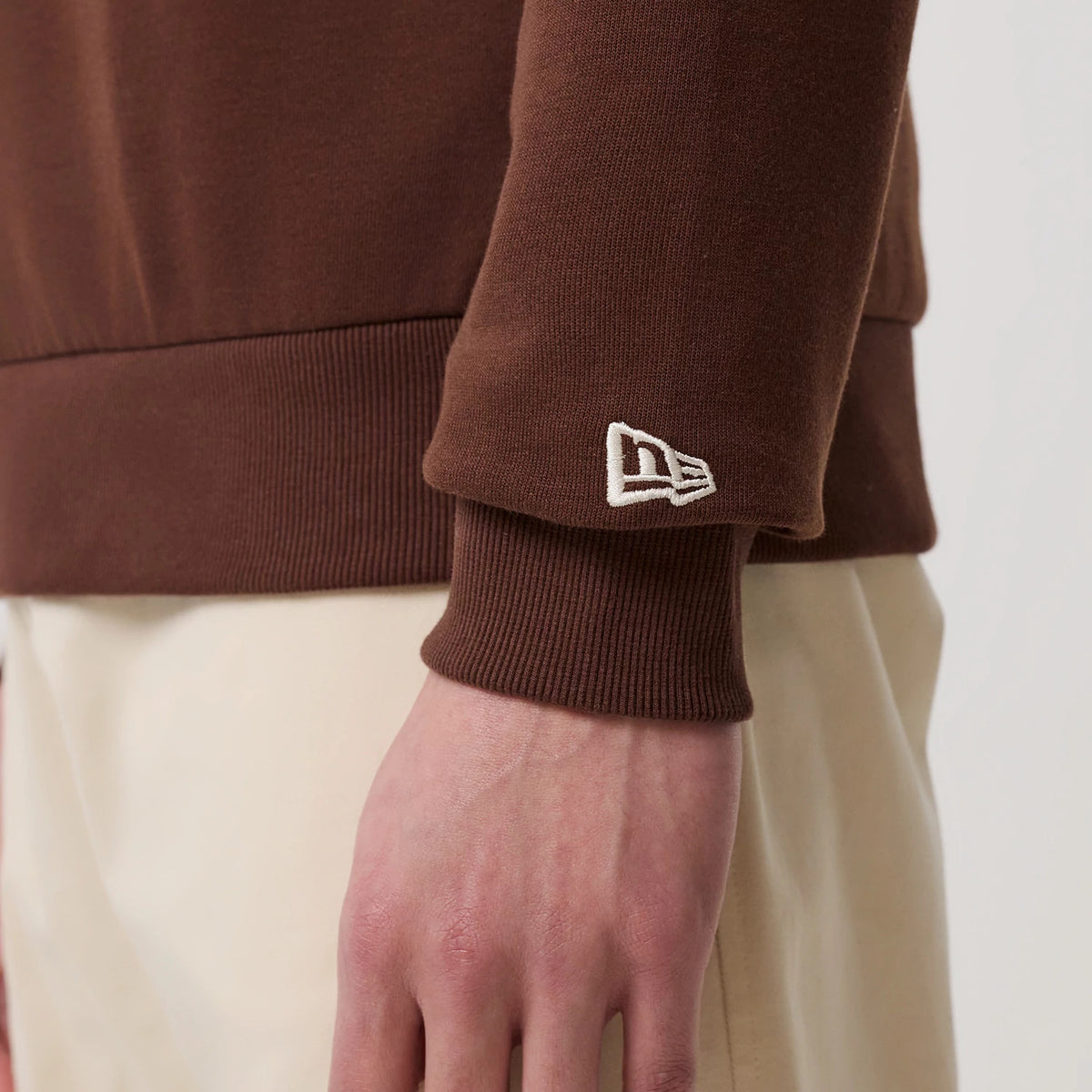 New Era LA Dodgers MLB Linear Brown Crew Neck Sweater, close-up view on a model showing the embroidered New Era flag logo on the left sleeve cuff, ribbed texture at the wrist, and brown fabric contrasting with light-coloured trousers.