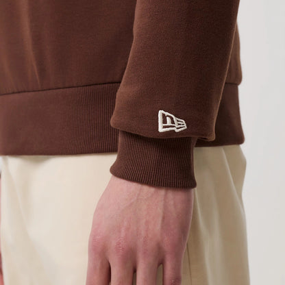 New Era LA Dodgers MLB Linear Brown Crew Neck Sweater, close-up view on a model showing the embroidered New Era flag logo on the left sleeve cuff, ribbed texture at the wrist, and brown fabric contrasting with light-coloured trousers.