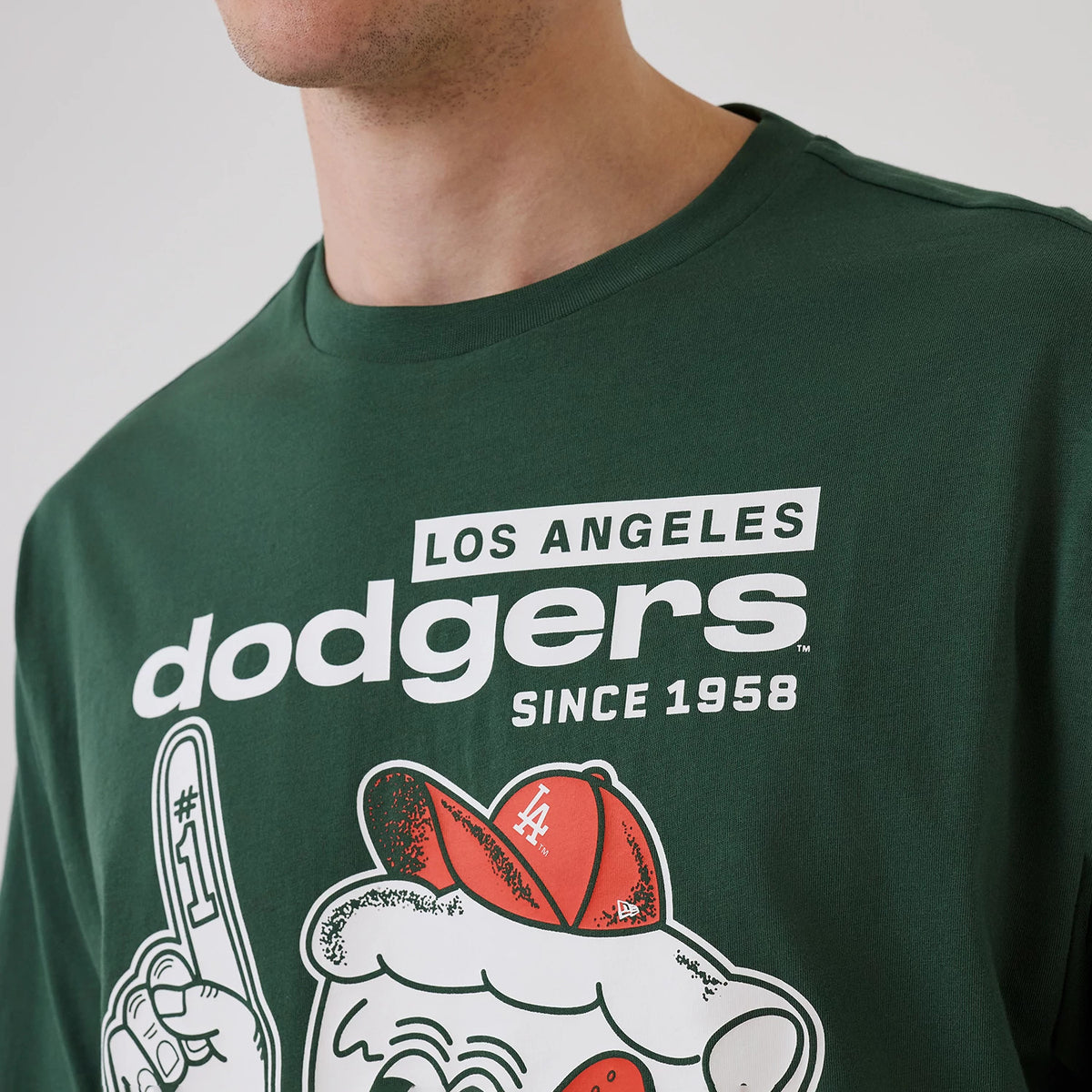 New Era LA Dodgers MLB Food Graphic Dark Green Oversized T-Shirt, front view on-model featuring prominent white Los Angeles Dodgers lettering and 'Since 1958', with a playful graphic of a character wearing a red cap with LA on its front.